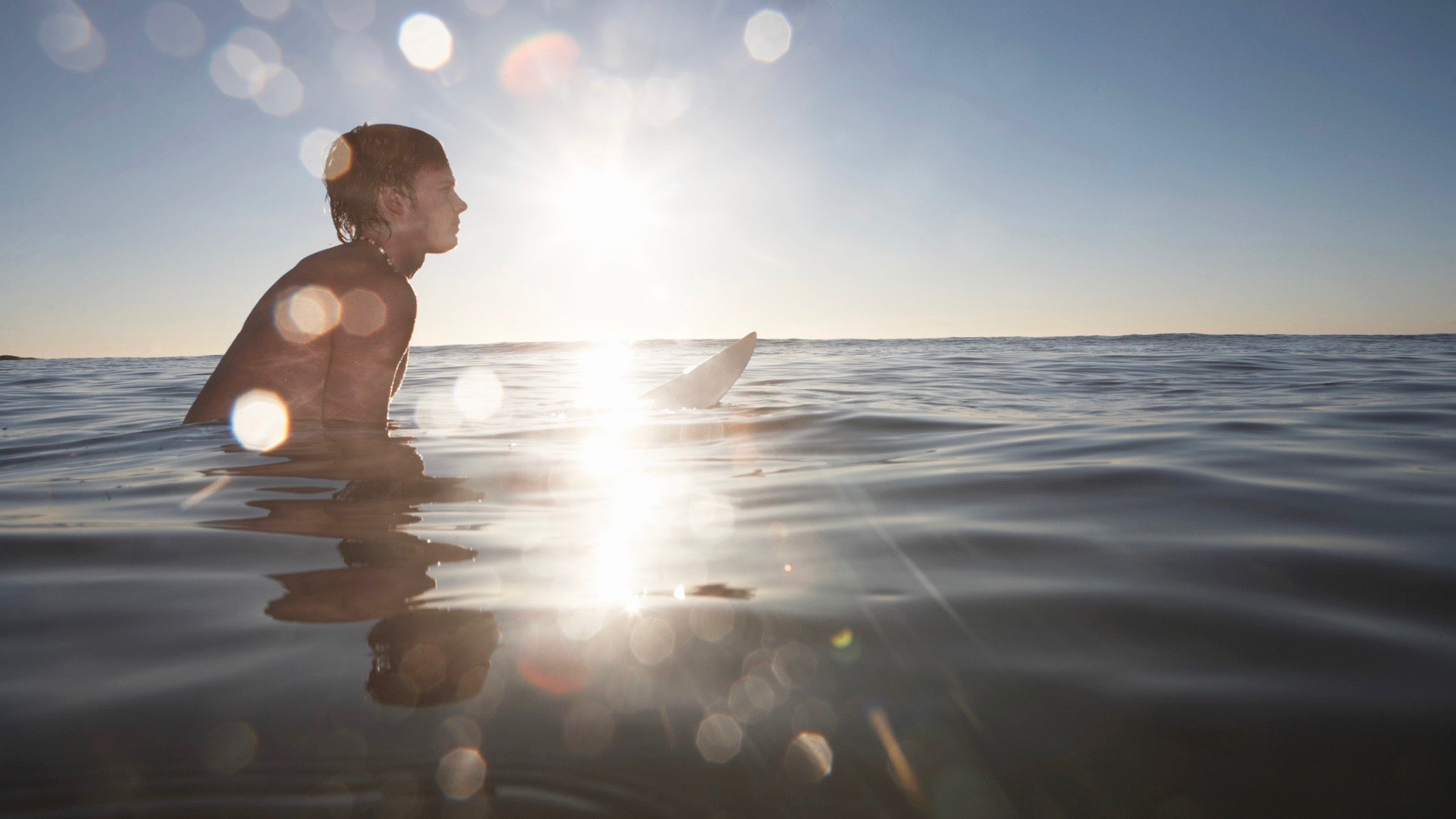 Teenage boy (15-17) in sea sitting on surfboard (sunflare)