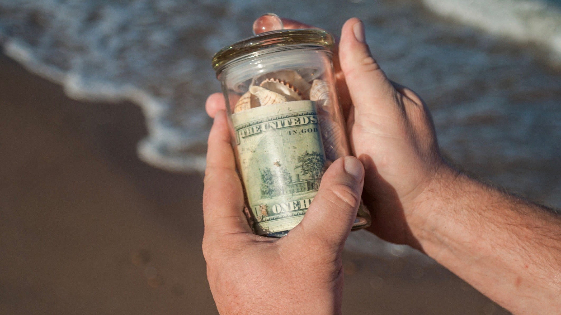 male hands holding a glass jar with money and shells found on the shore of the sea