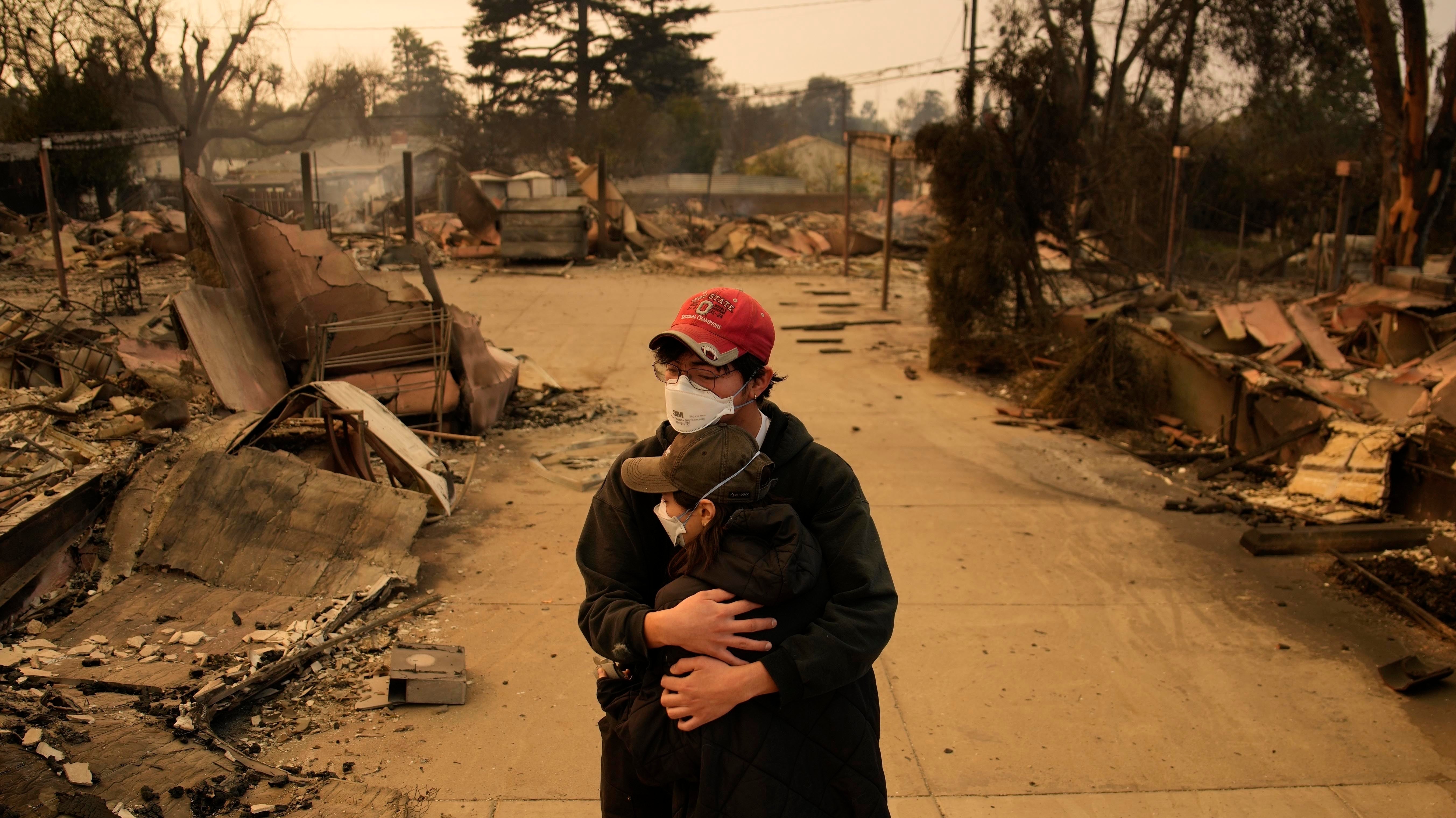 Two people embrace in front of wildfire ruins.