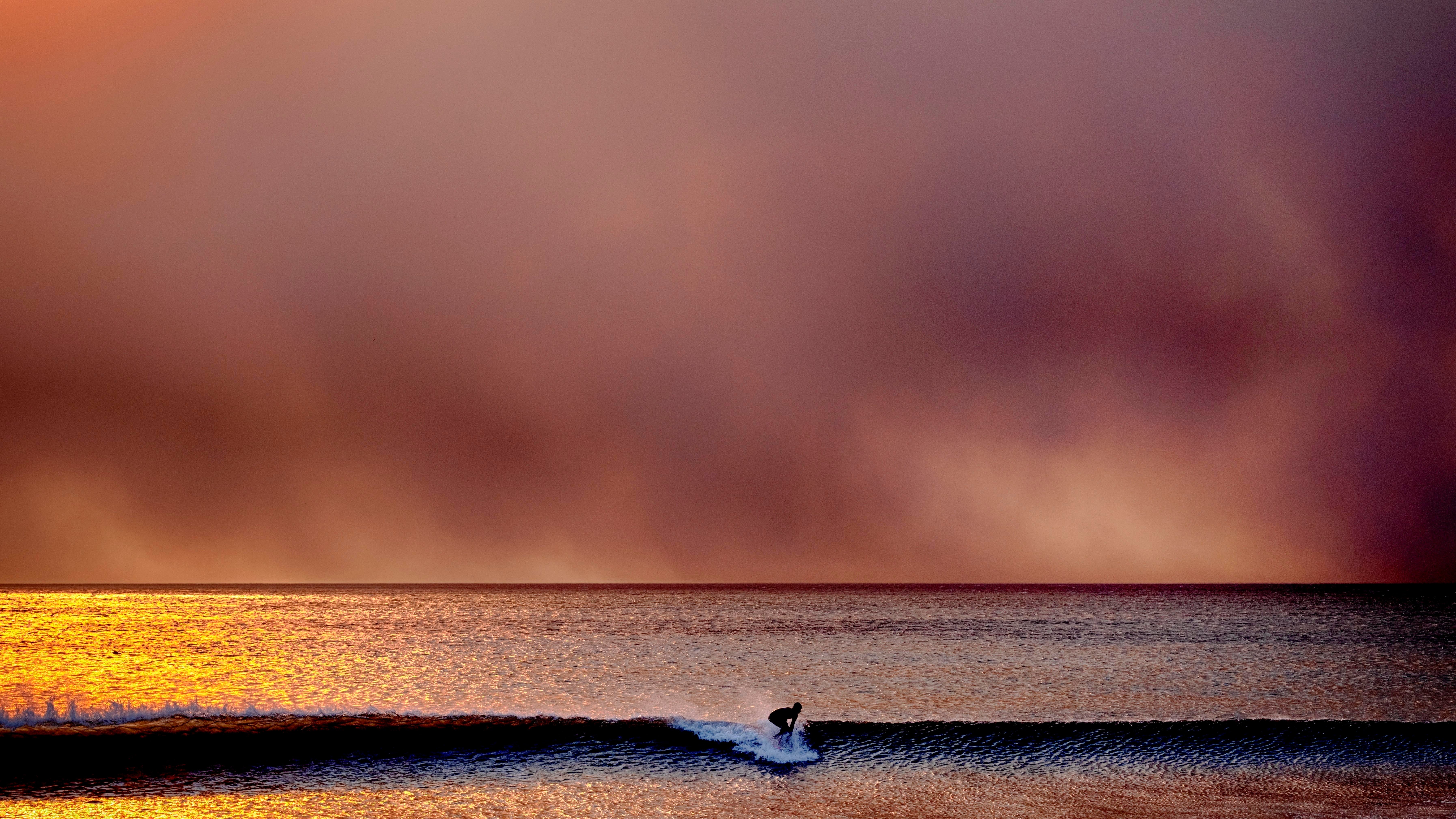 A surfer rides a wave against a backdrop of a red sky filled with smoke.