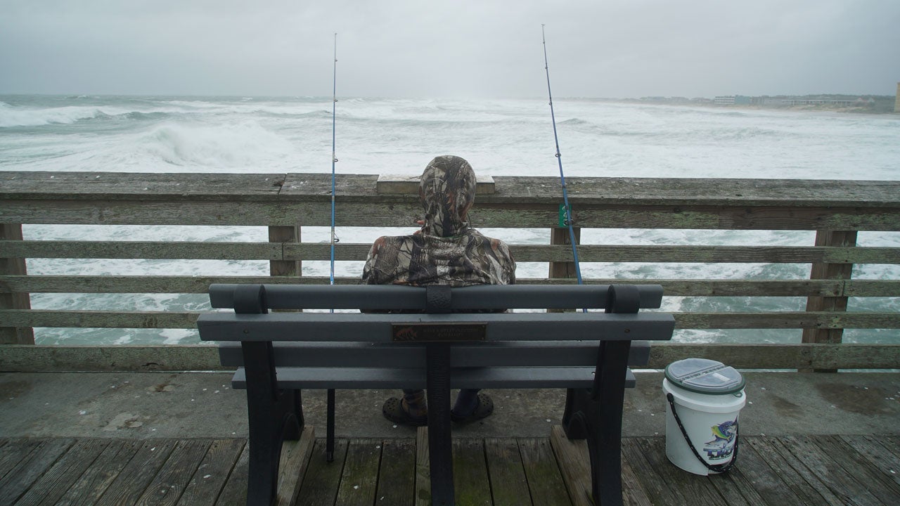 A man sits on a bench on a dock with fishing lures to his left and right, looking at the ocean