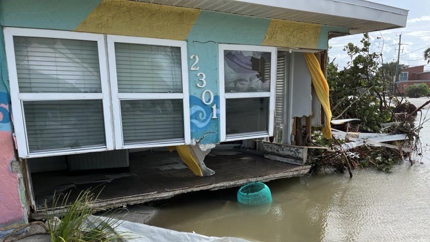 A house is shown surrounded by water with part of its lower wall washed out.