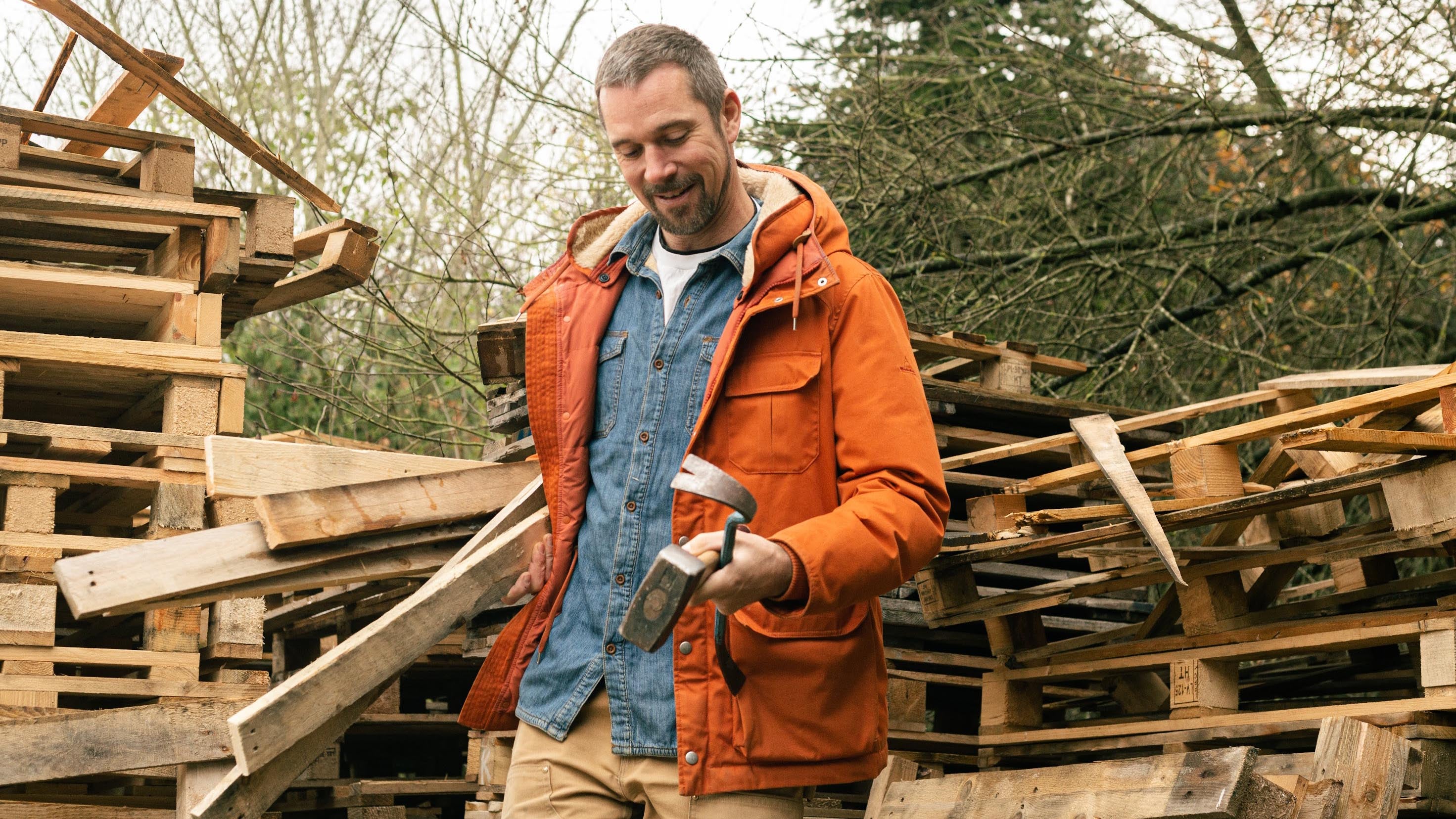 The artist in an orange jacket stands next to piles of pallets.
