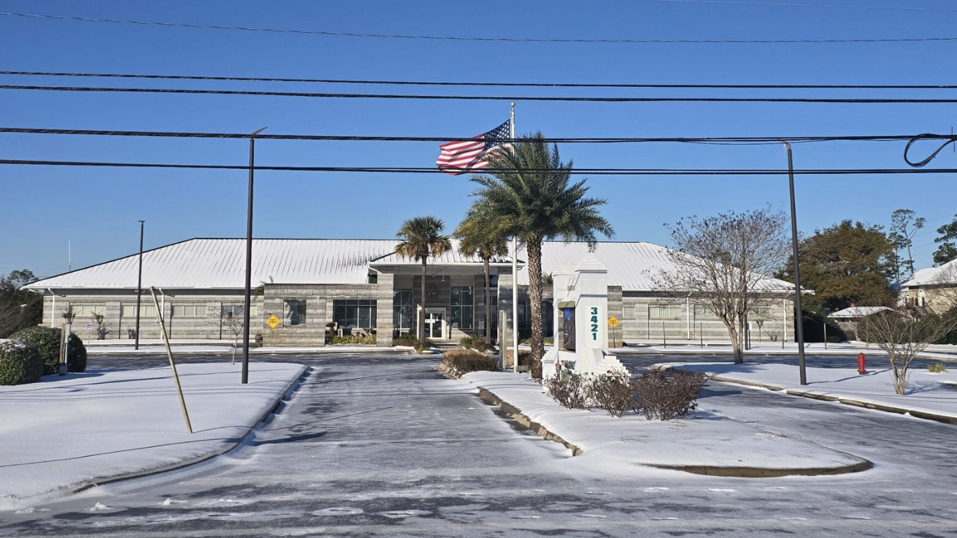 Snow covers the ground around palm trees and a single-story office building with the U.S. flag flying in front, against a brilliant blue sky..