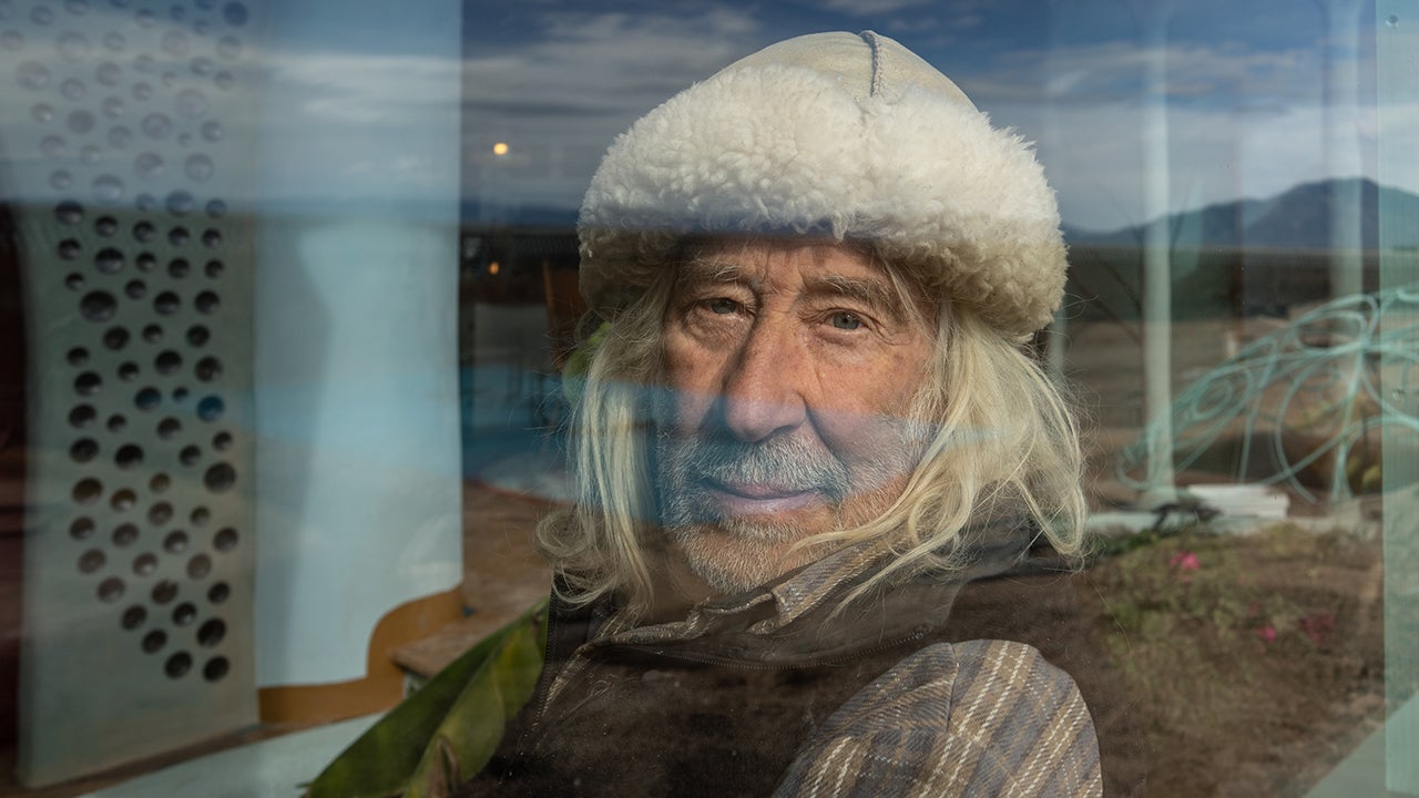 Michael Reynolds, an older gentleman with white hair, wearing a white, furry hat, sits in front of a window. 