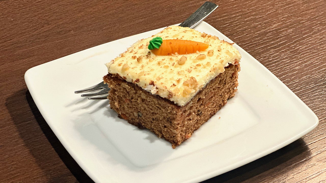A piece of carrot cake sits on a white plate with a fork with white icing, nuts and a piped carrot decoration on top. 
