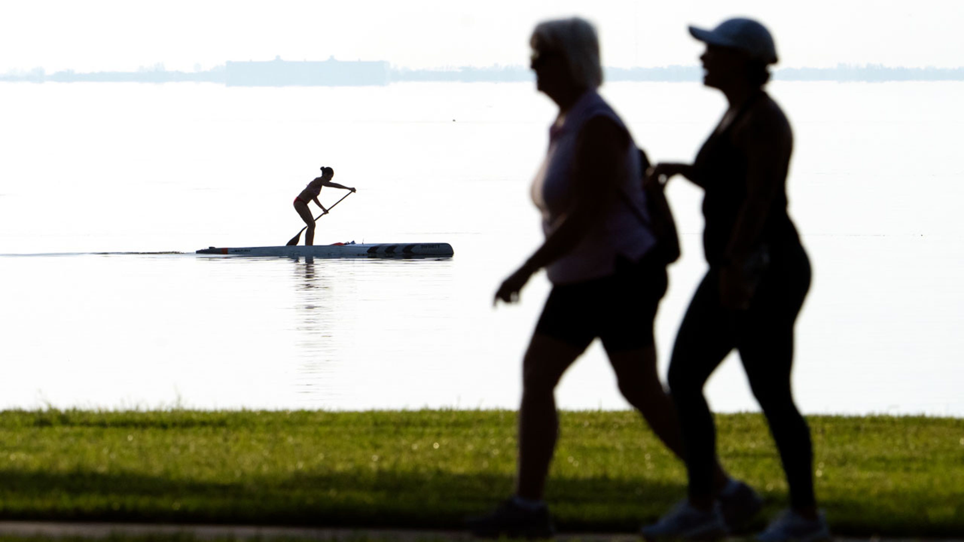 A person paddleboards across water as two people walk in the foreground
