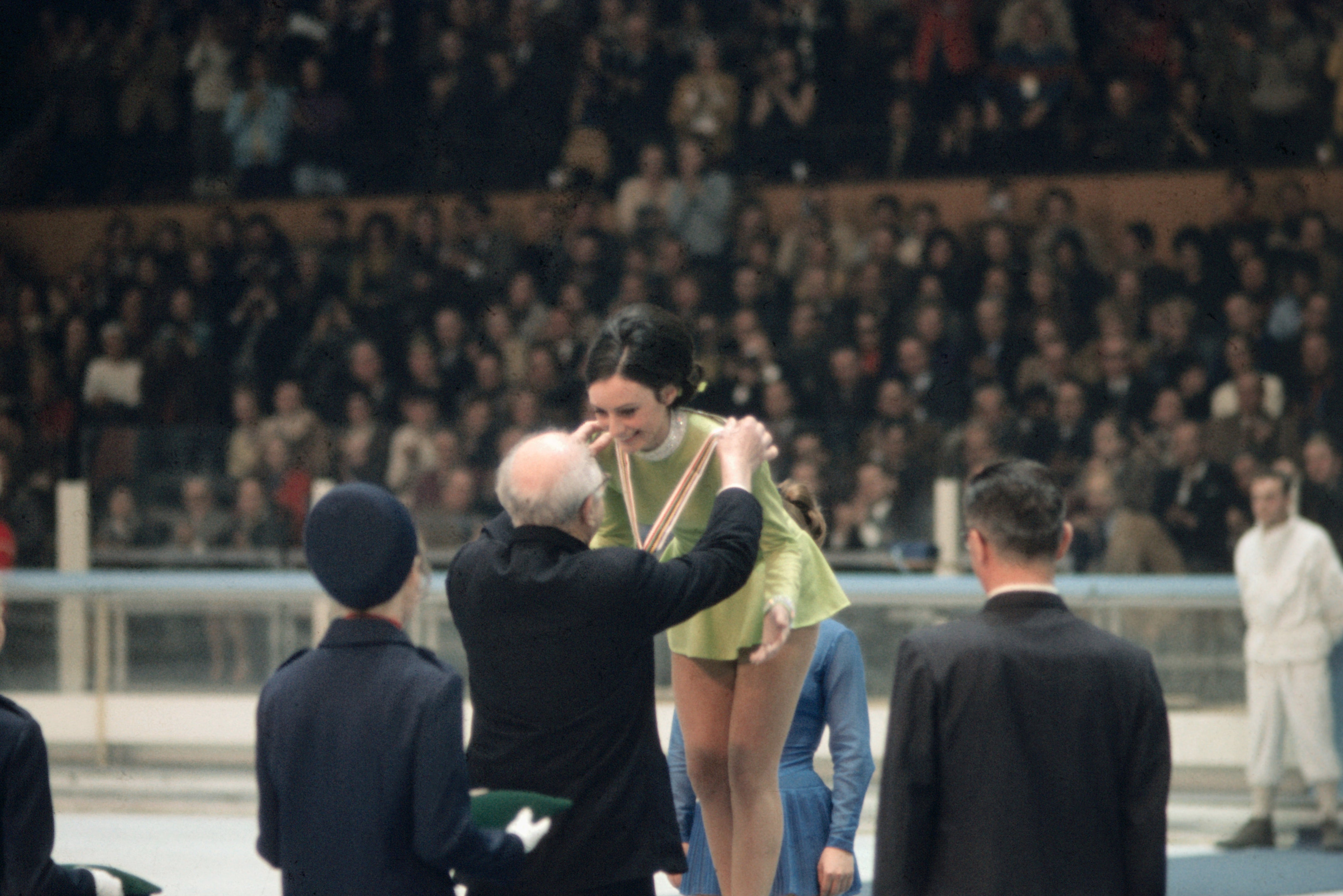 Peggy Fleming gracefully executing a spiral on the ice, wearing a white skating costume, during the 1968 Grenoble Winter Olympics.