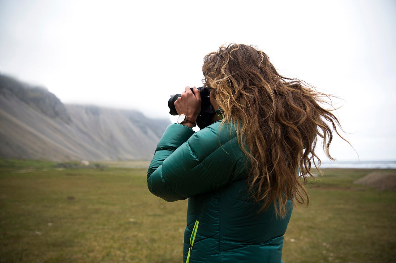 A woman in a green coat snaps a photo with mountains in the background.