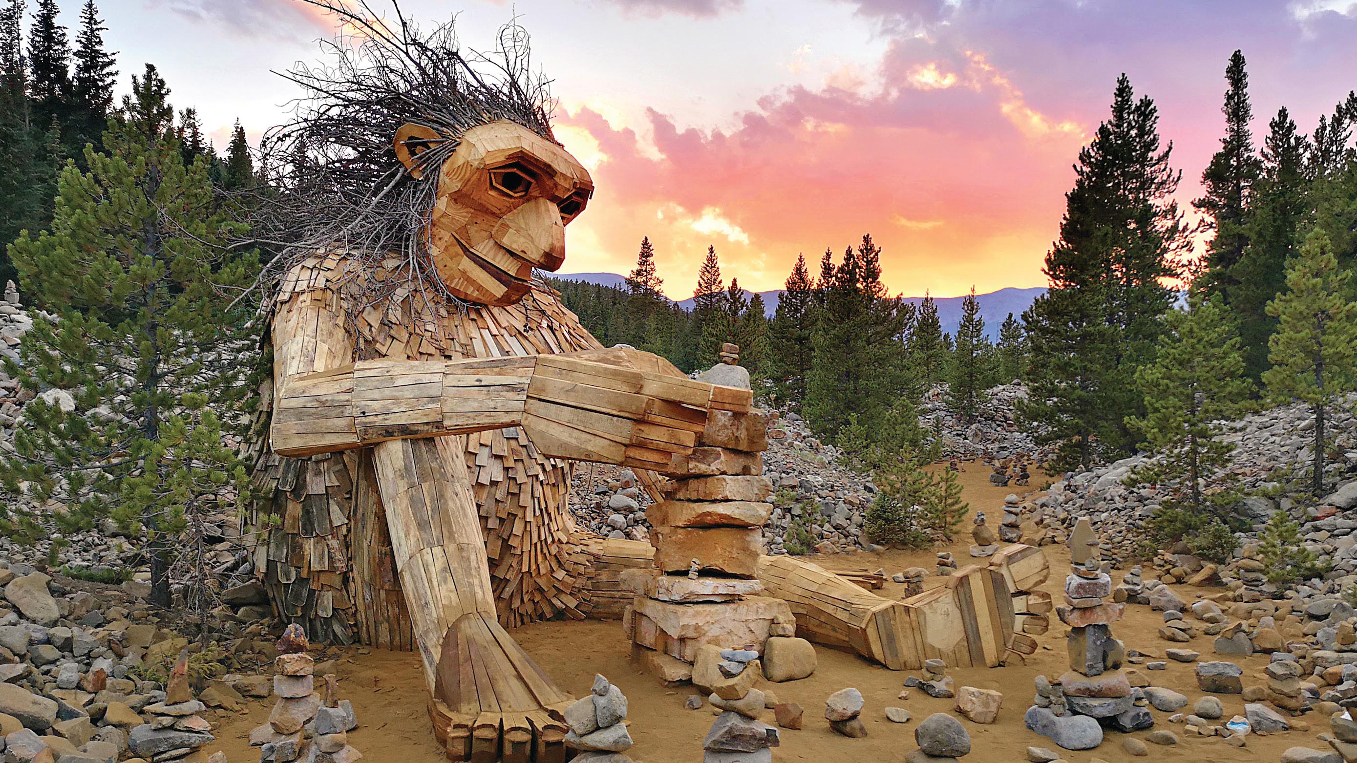 A giant troll made of wood sits taller than trees, with the sun setting in the background.