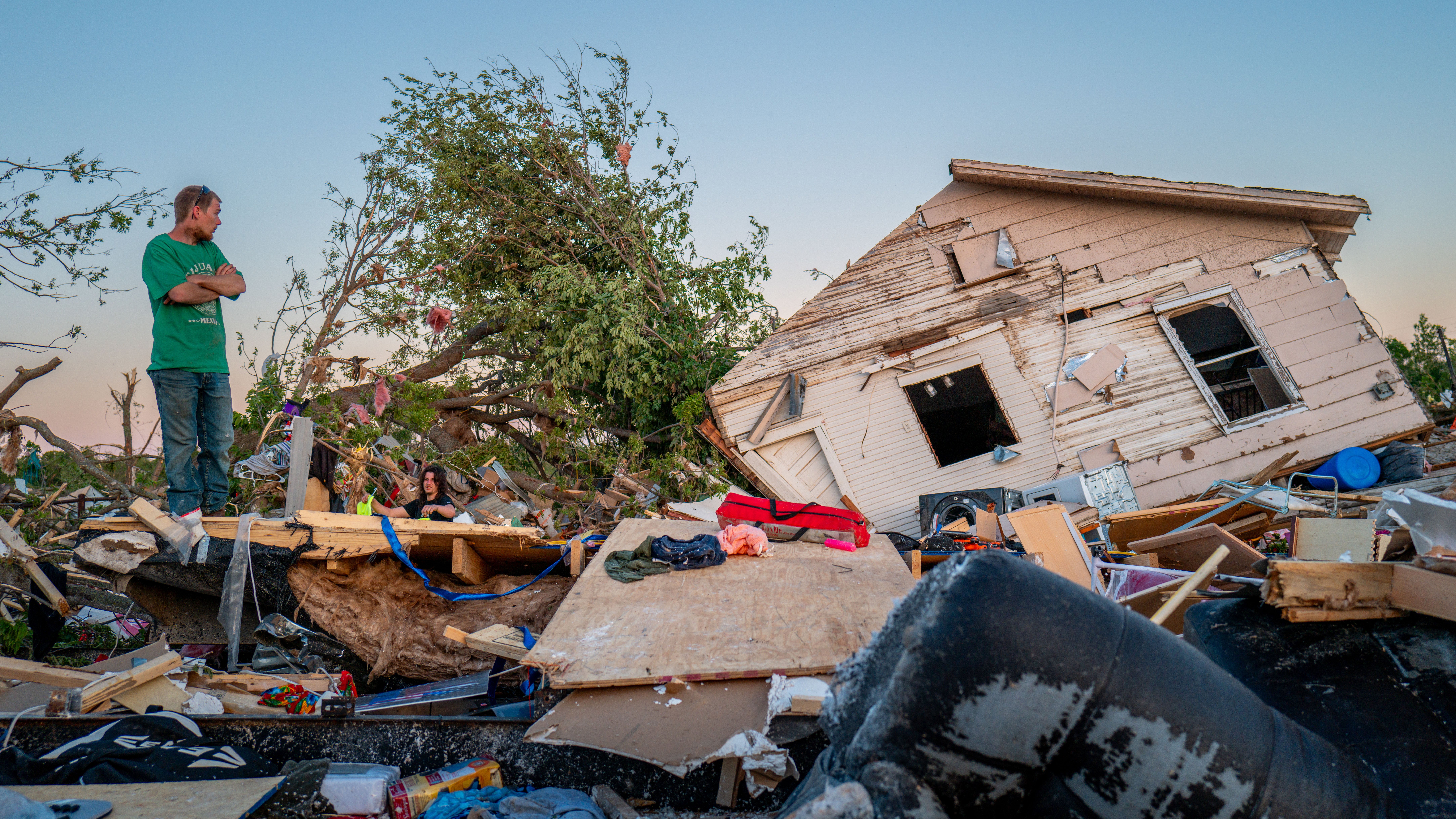 A man in a green shirt stands with arms folded on top of tornado damage next to a lopsided home.