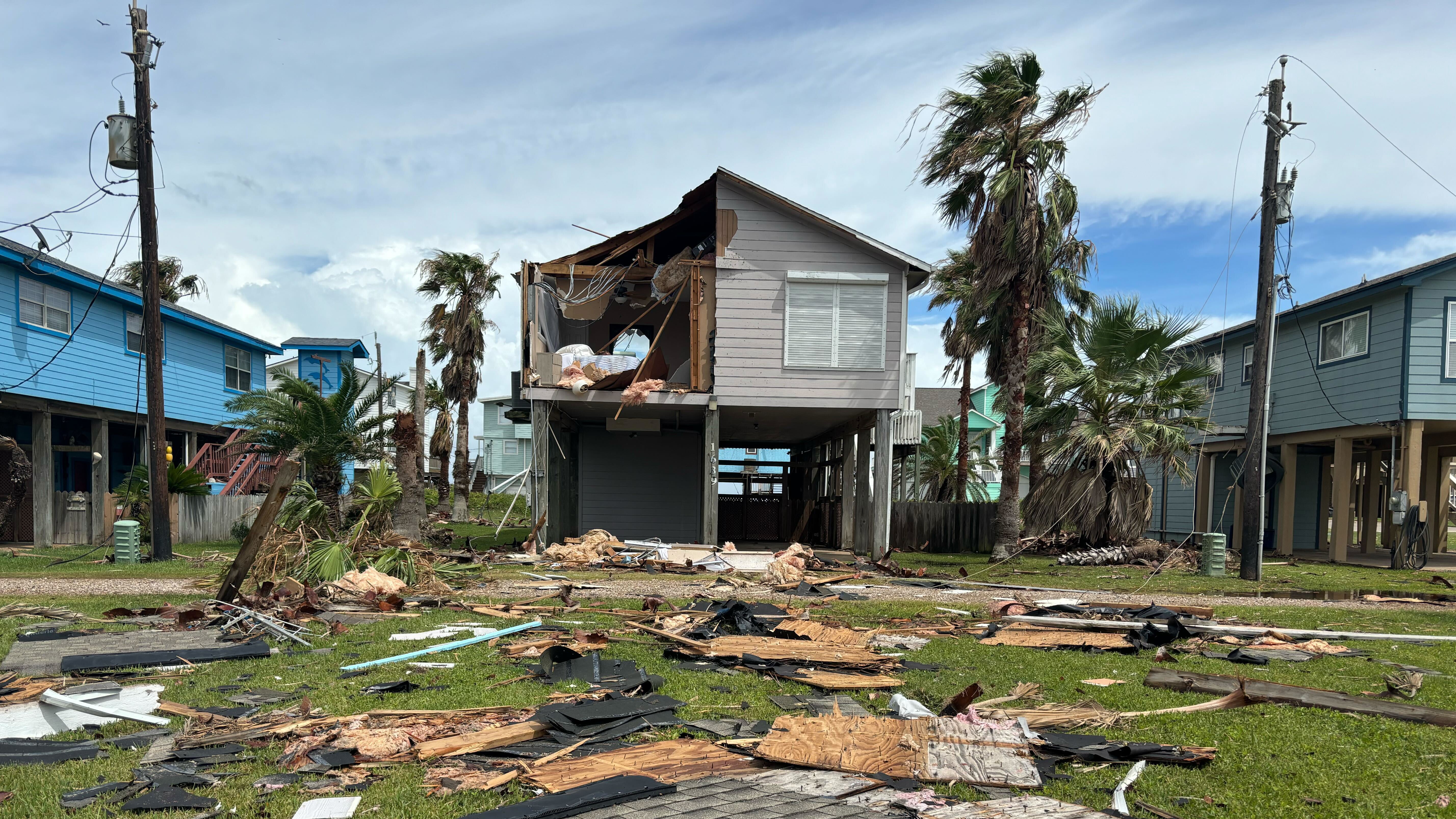 The top left corner of a home is torn off, revealing furniture and wires. Storm damage is scattered in the foreground.