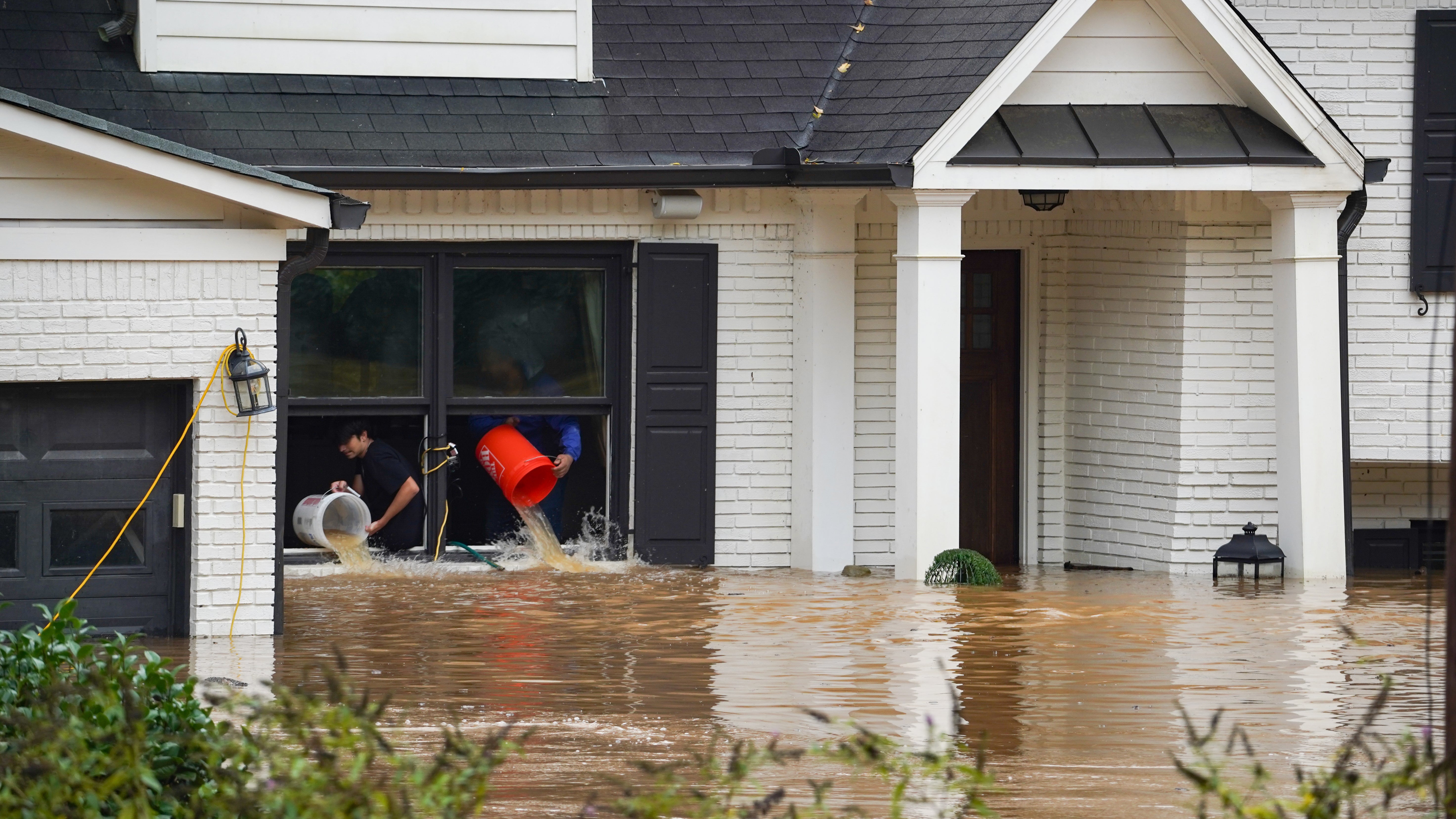 Floodwaters are seen up to the bottom of a first floor window of a home. Residents are bailing the home out with pails and throwing it out the window.