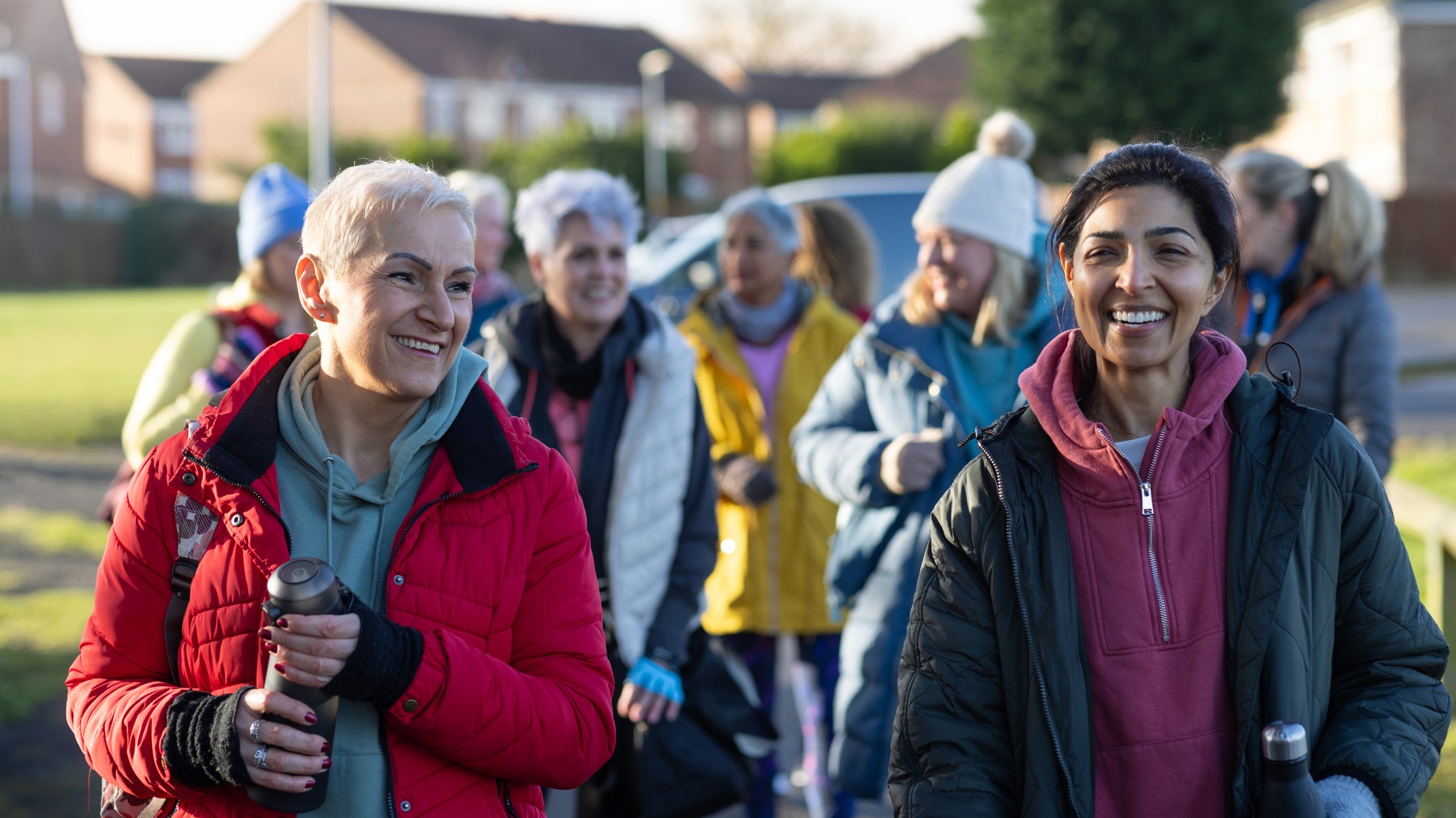 A waist-up shot of a group of mature women dressed in warm, sporty attire arriving to netball training together. They are smiling and carrying water bottles. Located in a suburban park area, with houses and greenery in the background