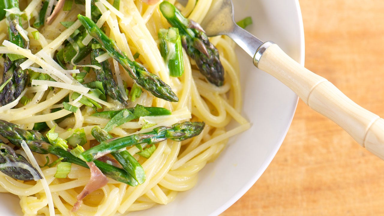 White bowl of creamy pasta primavera with asparagus, prosciutto, fresh herbs, parmesan cheese and freshly ground pepper from above.