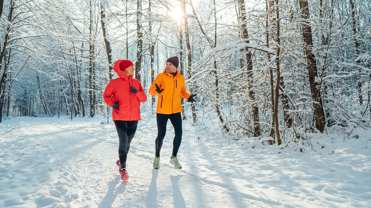 Middle-aged smiling trail couple runners man and woman dressed bright windproof jackets running speaking picturesque snowy forest during sunny frosty day. Sporty active people, winter training image.