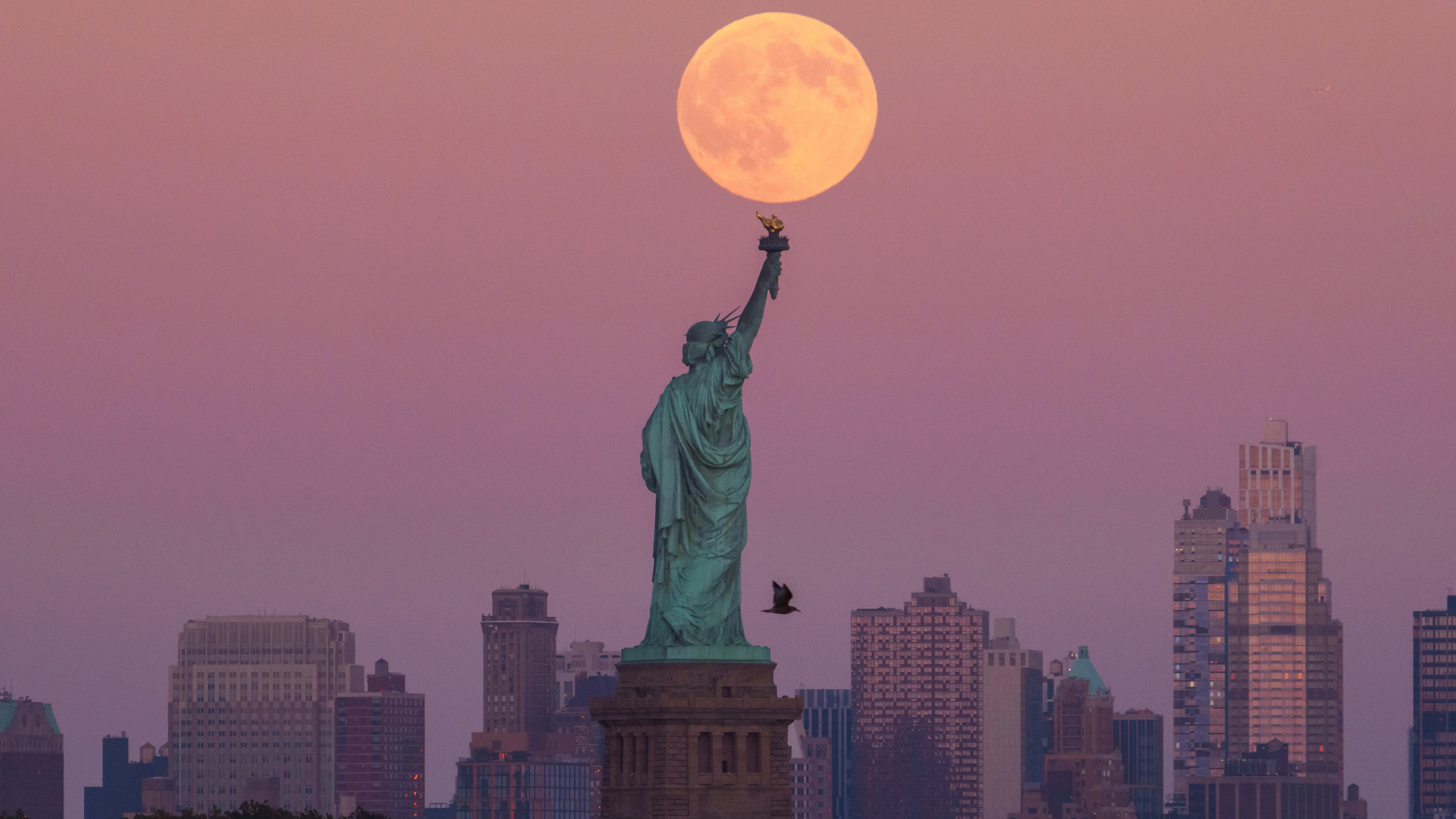The Harvest Supermoon rises behind the Statue of Liberty and the Brooklyn skyline.
