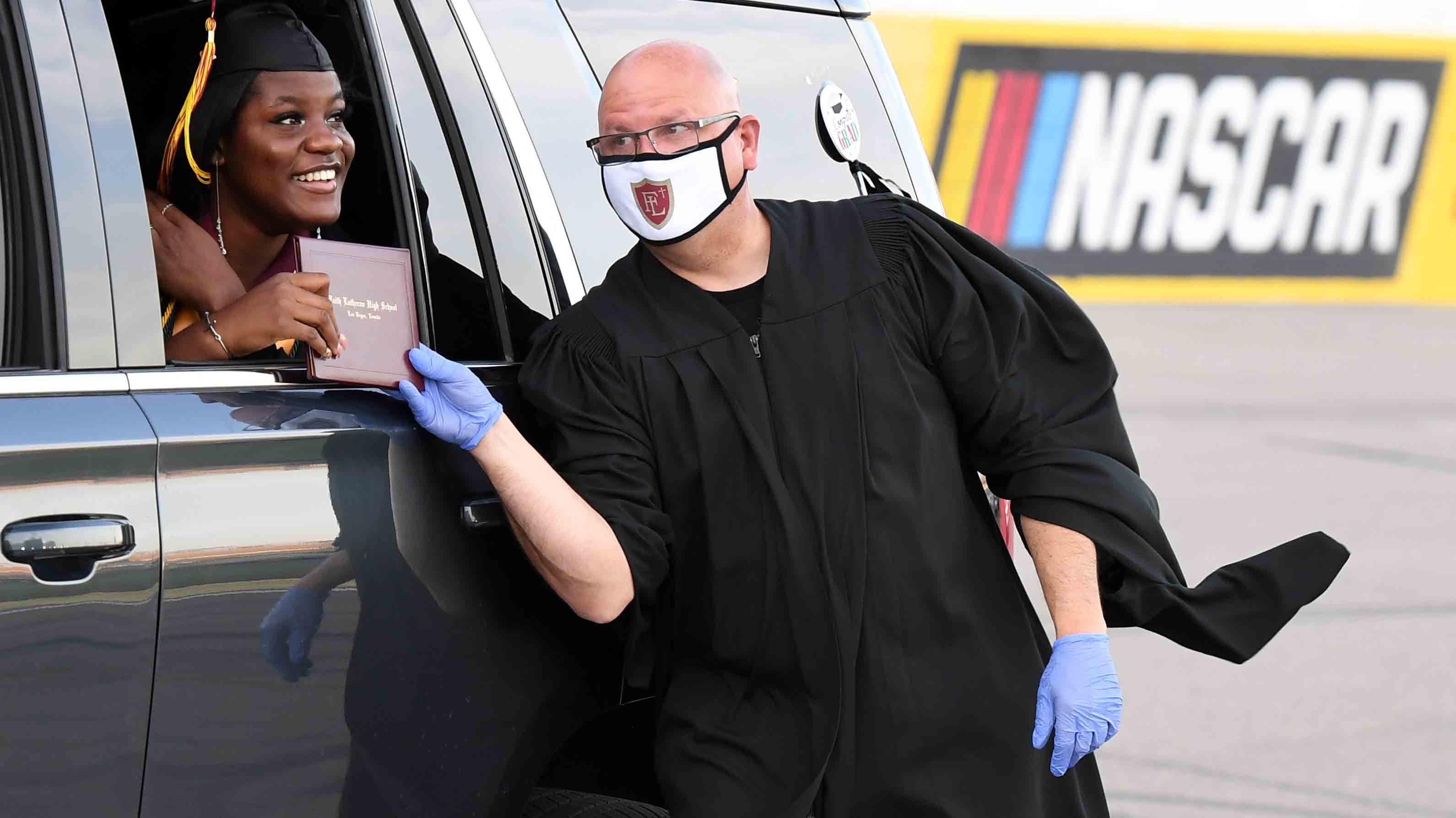 LAS VEGAS, NEVADA - MAY 22:  Faith Lutheran High School principal Scott Fogo (R) presents a graduate with her diploma during a commencement ceremony for Faith Lutheran held at Las Vegas Motor Speedway due to the spread of the coronavirus on May 22, 2020 in Las Vegas, Nevada. The graduation comes after the COVID-19 pandemic forced the closure of schools in Nevada for the remainder of the year. Instead of a traditional graduation, students received their diplomas as they drove across the start-finish line of the racetrack and then took about a one-mile victory lap before exiting the speedway. The students and their families could listen to the commencement address and other remarks from the valedictorian on their vehicle radios via the speedway's FM transmitter.  (Photo by Ethan Miller/Getty Images)