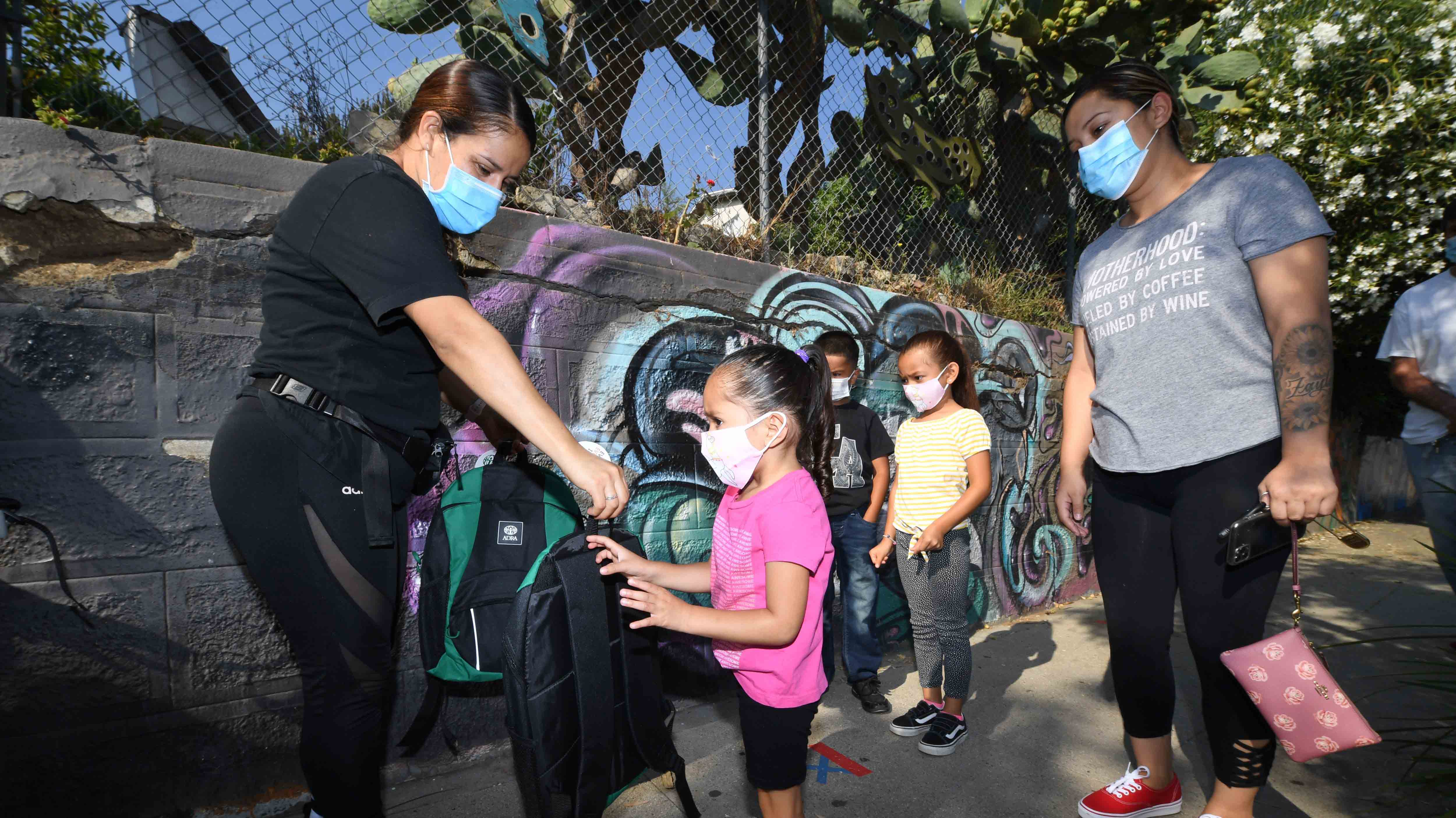 A volunteer (L) hands three-year-old Zayla a backpack filled with school supplies at a distribution to support neighborhood families as her mom Claudia Rodriguez (R) looks on, August 14, 2020 in Los Angeles, California. - The back-to-school giveaway is being held as part of Adventist Health White Memorial hospital's series of COVID-19 community rescue programs.  As the coronavirus pandemic continues all  Los Angeles Unified School District (LAUSD) schools will be closed and students will return to class via remote learning from home when the 2020-21 school year starts on August 18, 2020. (Photo by Robyn Beck / AFP) (Photo by ROBYN BECK/AFP via Getty Images)