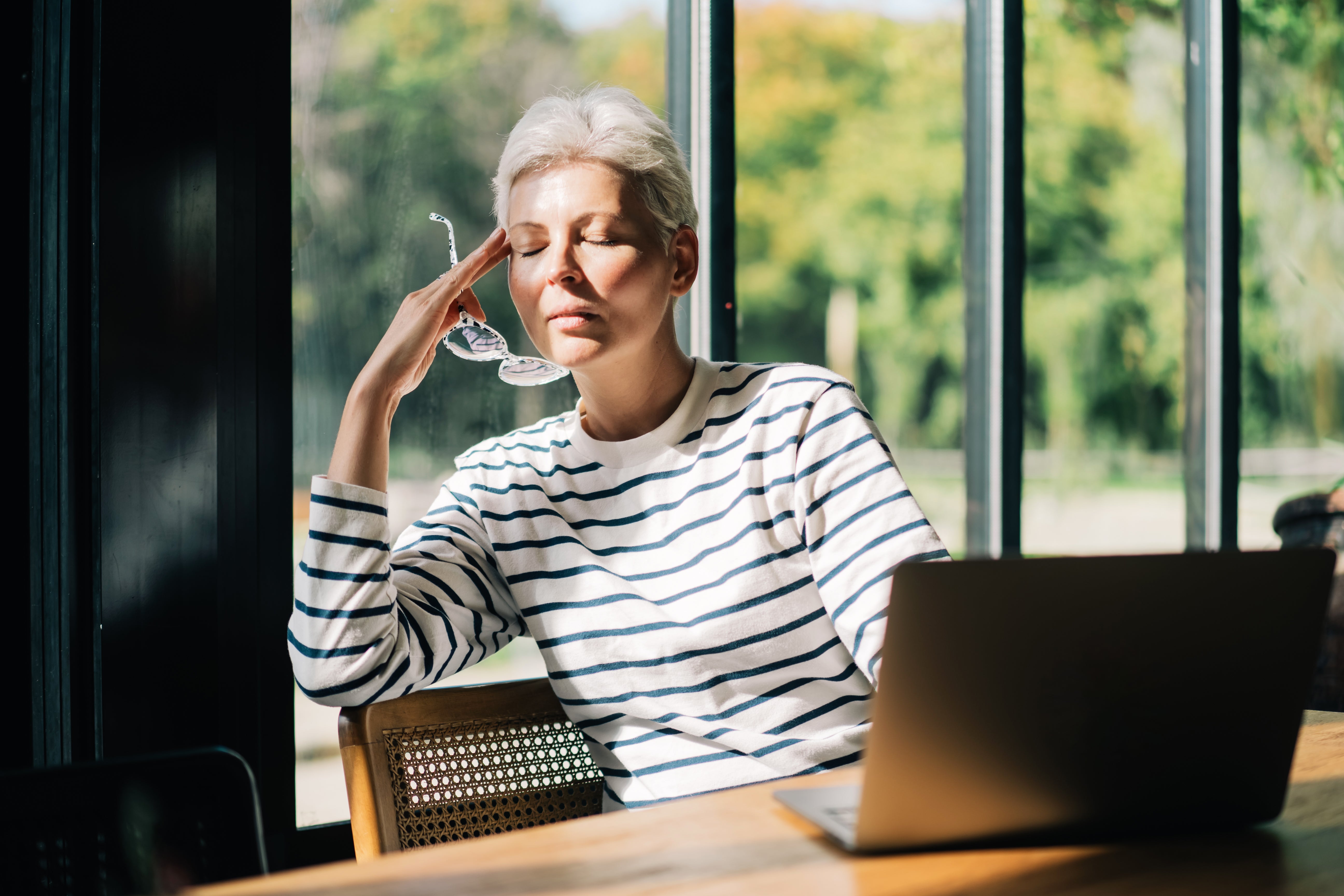 A middle-aged woman with short grey hair, wearing glasses and striped long-sleeve, is working with a laptop at her cafe. She&rsquo;s taken her glasses off, rubbing the temples with eyes closed, experiencing a headache. Silver generation business owners feeling stress, conquering adversities. She is positive about future and utilizing innovation to cope with financial crisis