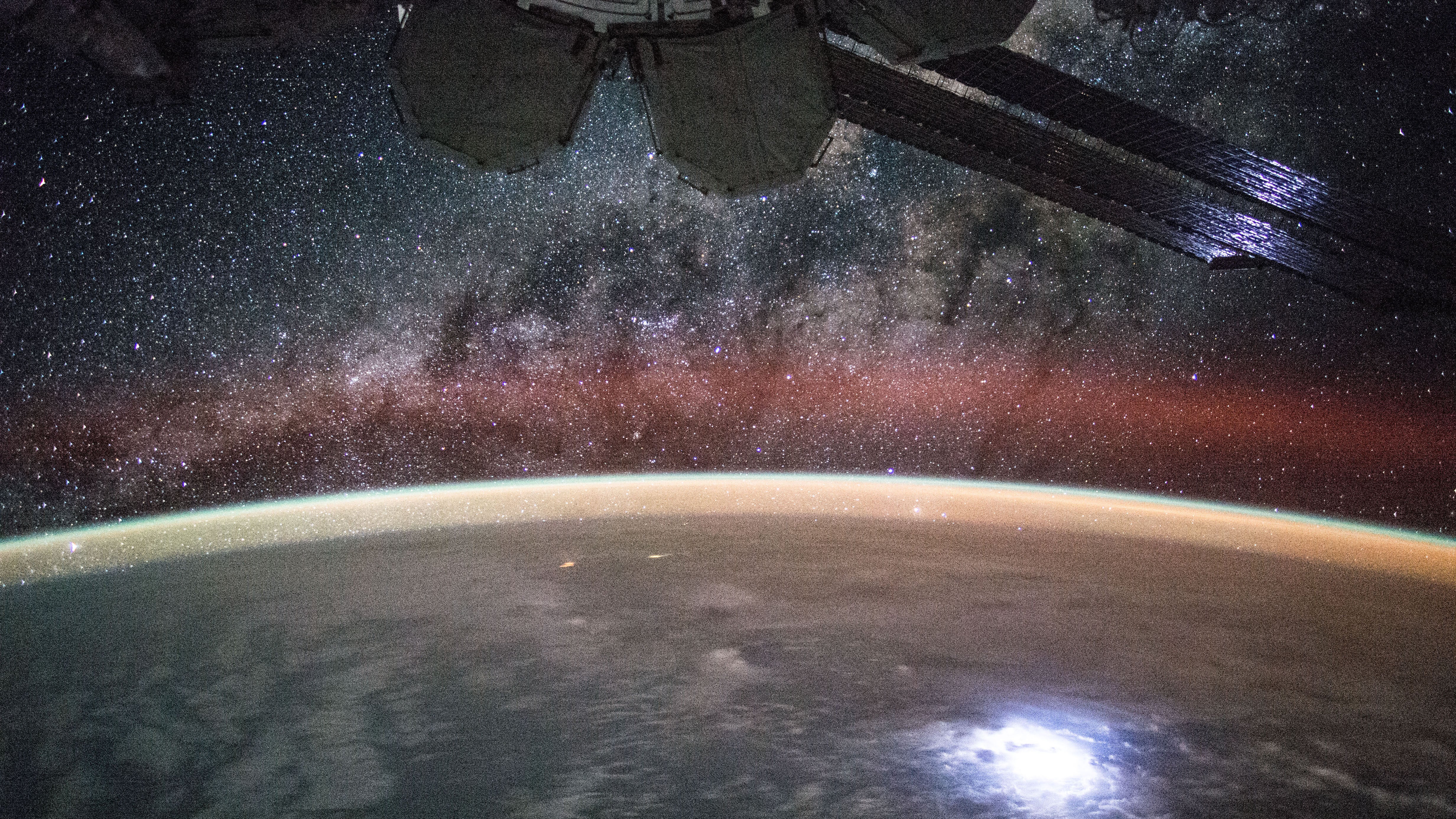 How many of these can you find in this photograph: an aurora, airglow, one of the oldest impact craters on the Earth, snow and ice, stars, city lights, and part of the International Space Station? Most of these can be identified by their distinctive colors. The aurora here appears green at the bottom, red at the top, and is visible across the left of image. Airglow appears orange and can be seen hovering over the curve of the Earth. The circular Manicouagan Crater in Canada, about 100 kilometers across and 200 million years old, is visible toward the lower right and is covered in white snow and ice. Stars, light in color, dot the dark background of space. City lights appear a bright yellow and dot the landscape. Finally, across the top, part of the International Space Station (ISS) appears mostly tan. This image was taken from the ISS in 2012. (NASA)
