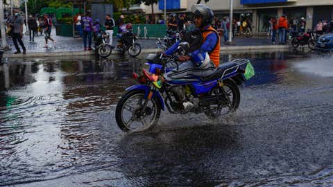 A man rides his motorcycle over a puddle in Caracas, Venezuela, Wednesday, June 29, 2022. As the latest tropical disturbance advances through the area, Venezuela shuttered schools, opened shelters and restricted air and water transportation on Wednesday as President Nicolas Maduro noted that the South American country already has been struggling with recent heavy rains. (AP Photo/Ariana Cubillos)