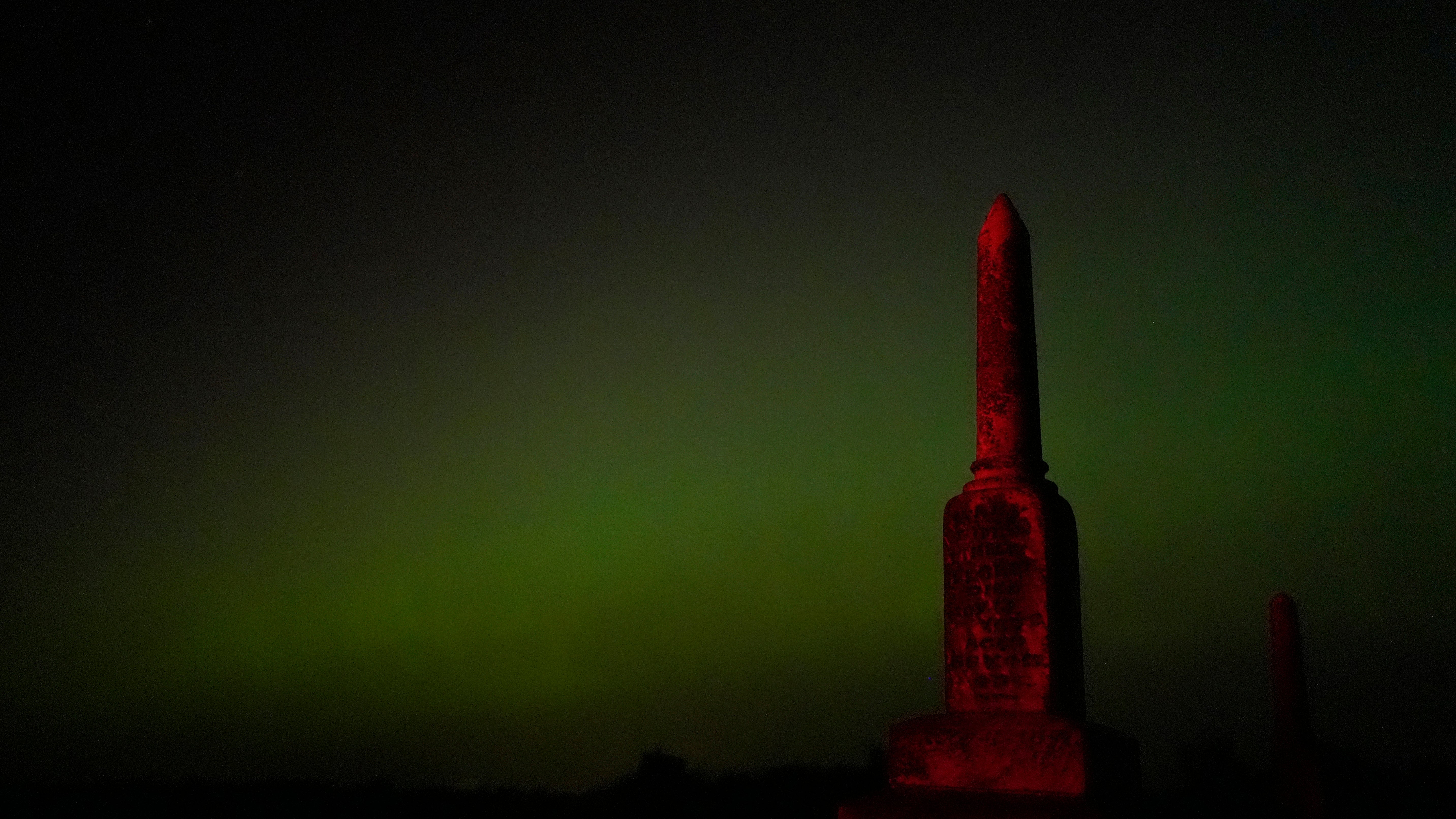 An old tombstone stands against the northern lights at a cemetery early Saturday, May 11, 2024, near Skidmore, Mo. (AP Photo/Charlie Riedel)