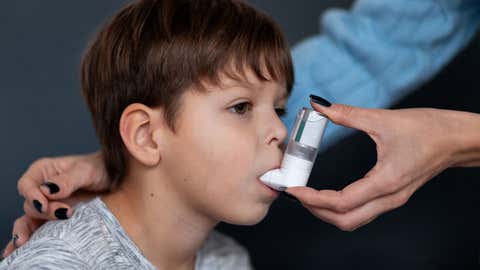 An adult's hands help a boy use an inhaler. The adult's hands gently embrace the child with care. The concept focuses on caring for children with chronic illnesses and home treatment.
