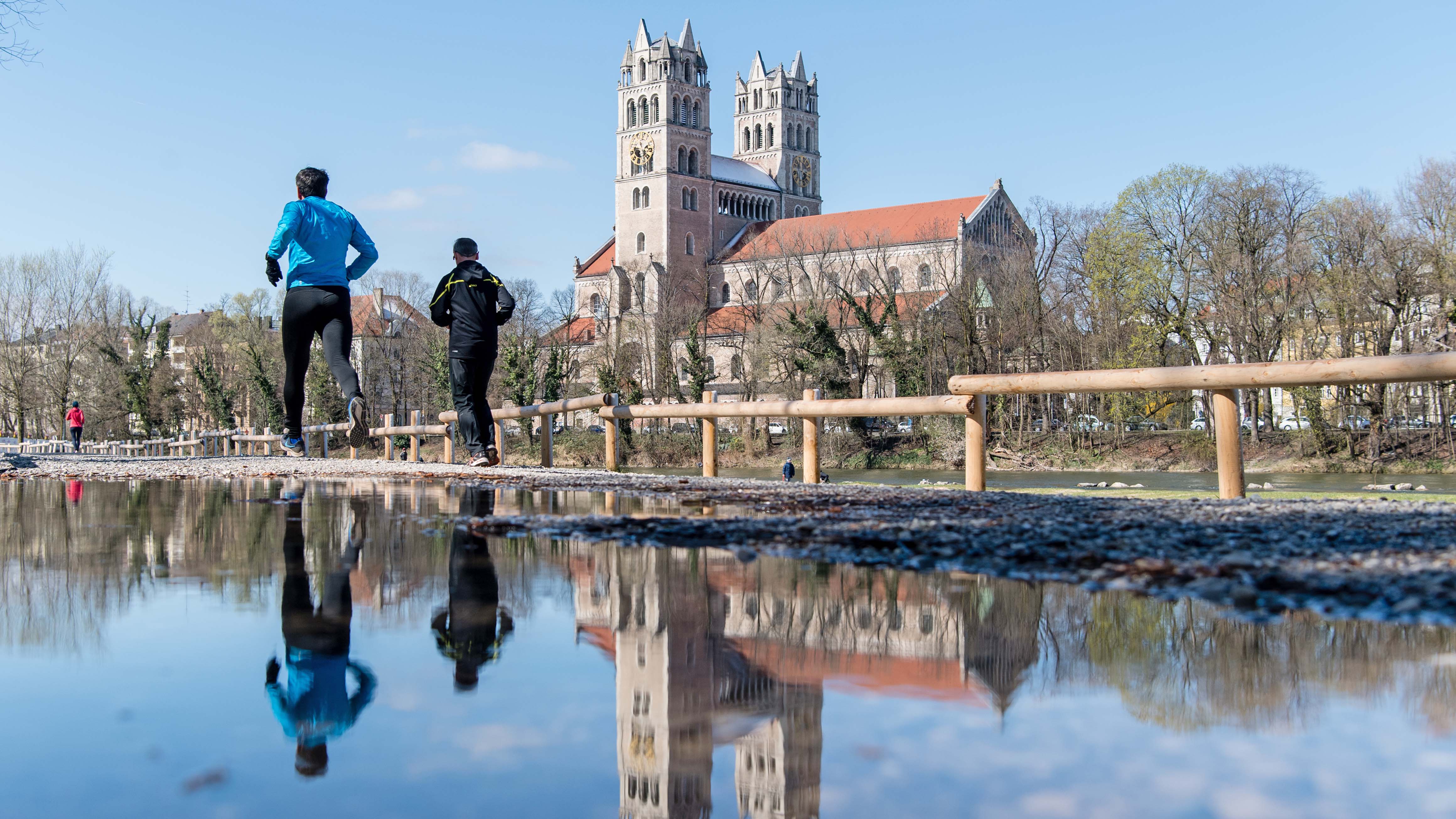 22.03.2020, Bayern, M&uuml;nchen: Zwei Jogger laufen an der Kirche St. Maximilian vor&uuml;ber. Das Motiv spiegelt sich dabei in einer gro&szlig;en Pf&uuml;tze. Trotz der neu auferlegten Ausgangsbeschr&auml;nkungen sind Sport an der freien Luft und Spazierg&auml;nge mit Personen des eigenen Haustands erlaubt. Foto: Matthias Balk/dpa +++ dpa-Bildfunk +++
