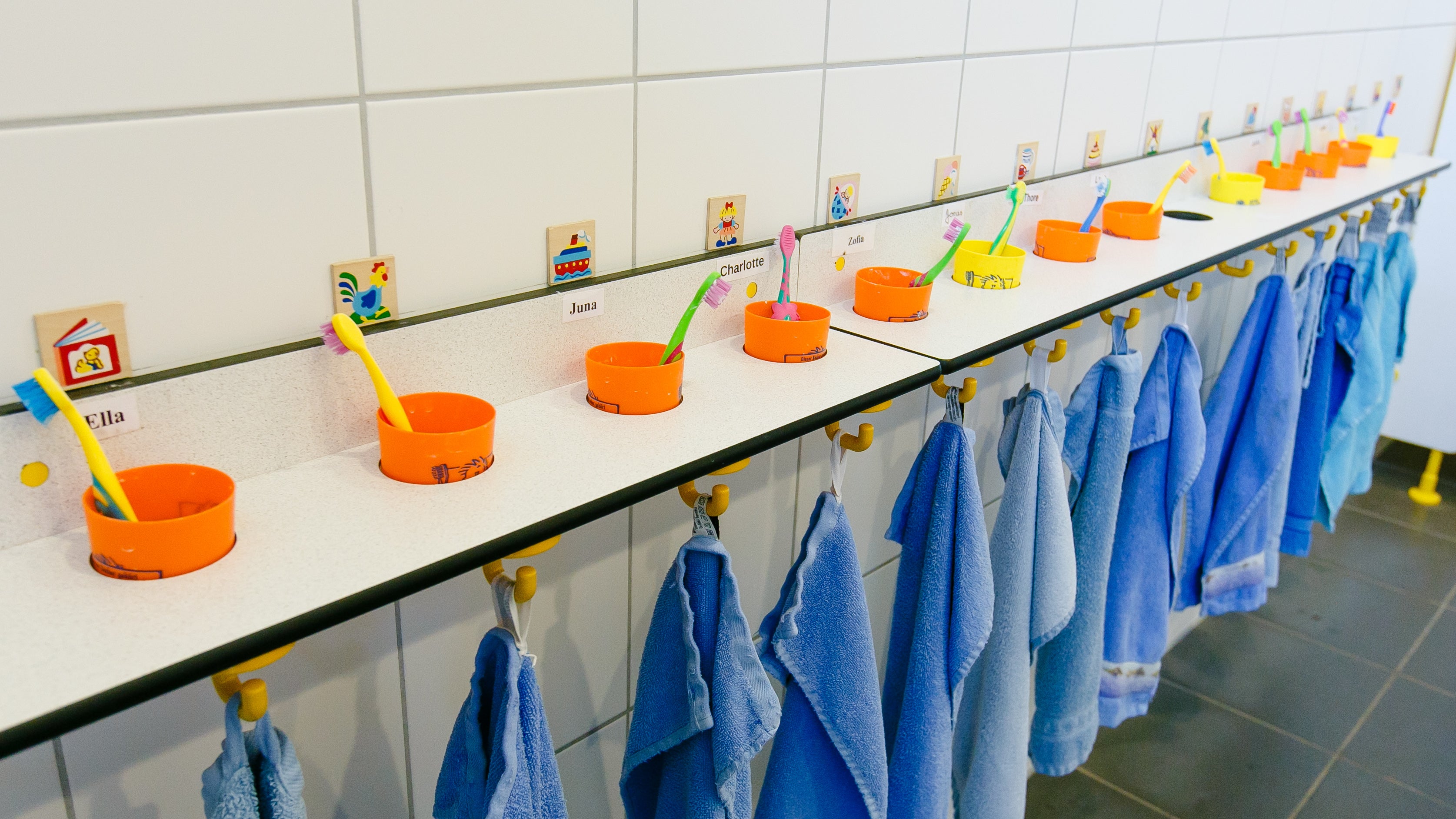 Zahnb&uuml;rsten in Zahnputzbechern stehen auf einem Regal &uuml;ber Handt&uuml;chern auf der Toilette einer Kindertagesst&auml;tte.