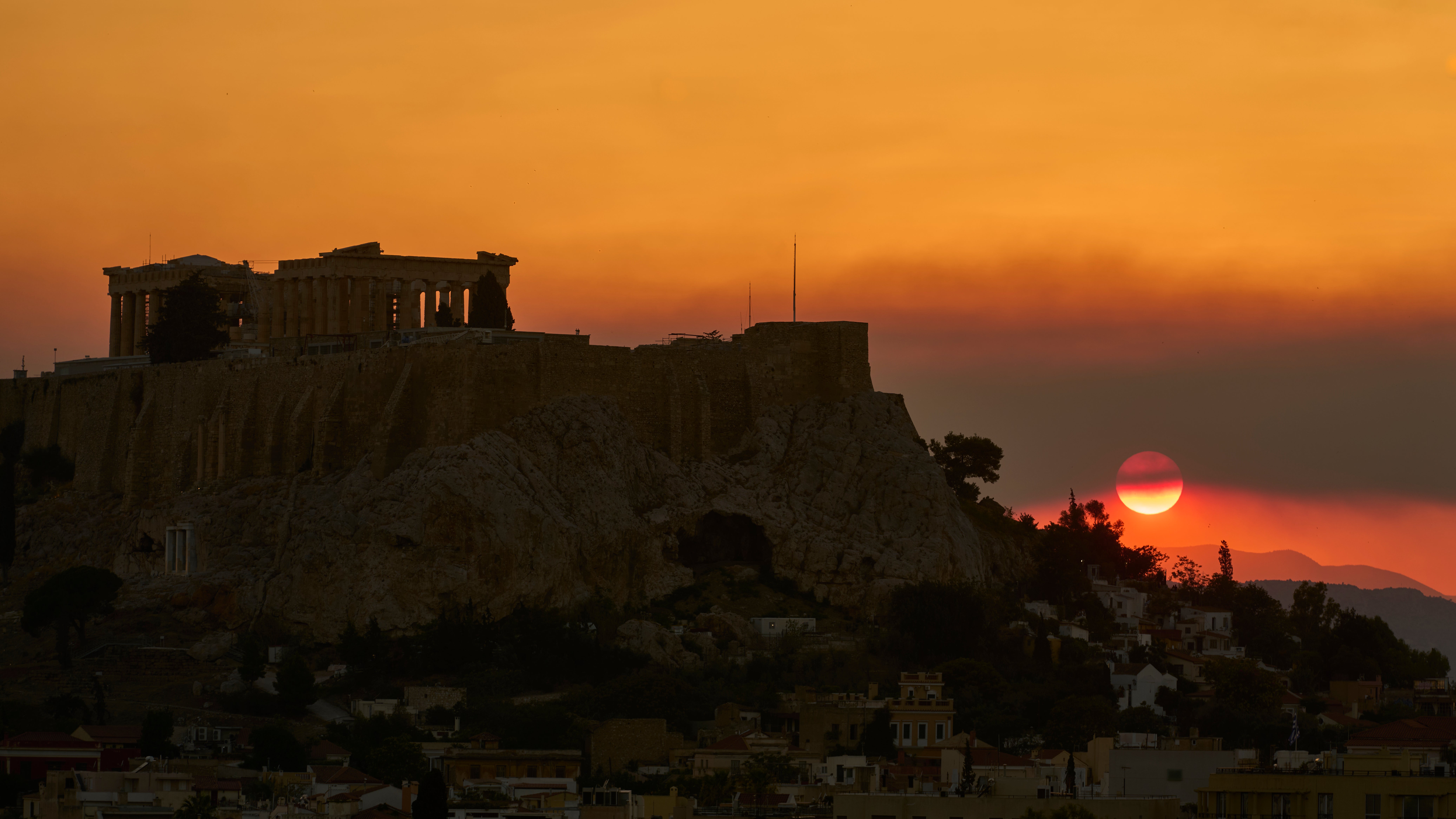 26.07.2025, Griechenland, Athen: Die Sonne geht hinter dem Akropolis-H&uuml;gel unter, w&auml;hrend der Rauch eines nahe gelegenen Waldbrandes den Himmel f&uuml;llt. (Petros Giannakouris/AP/dpa)
