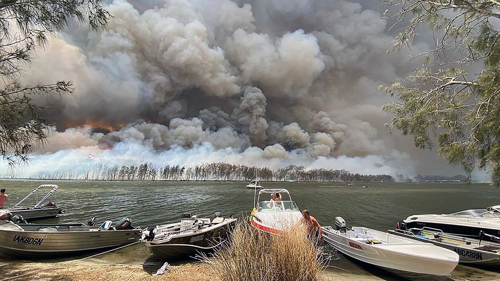 dpatopbilder - 02.01.2020, Australien, Lake Conjola: Boote werden an Land gezogen, w&auml;hrend im Hintergrund Rauchwolken &uuml;ber dem Ufer hinter dem Canjola See aufsteigen. Schon seit Oktober 2019 w&uuml;ten die Buschbr&auml;nde auf dem Kontinent. Allein in New South Wales ist mittlerweile etwa eine Fl&auml;che der Gr&ouml;&szlig;e Belgiens abgebrannt und landesweit wurden mehr als 1400 H&auml;user zerst&ouml;rt. Foto: Robert Oerlemans/Robert Oerlemans/AP/dpa +++ dpa-Bildfunk +++