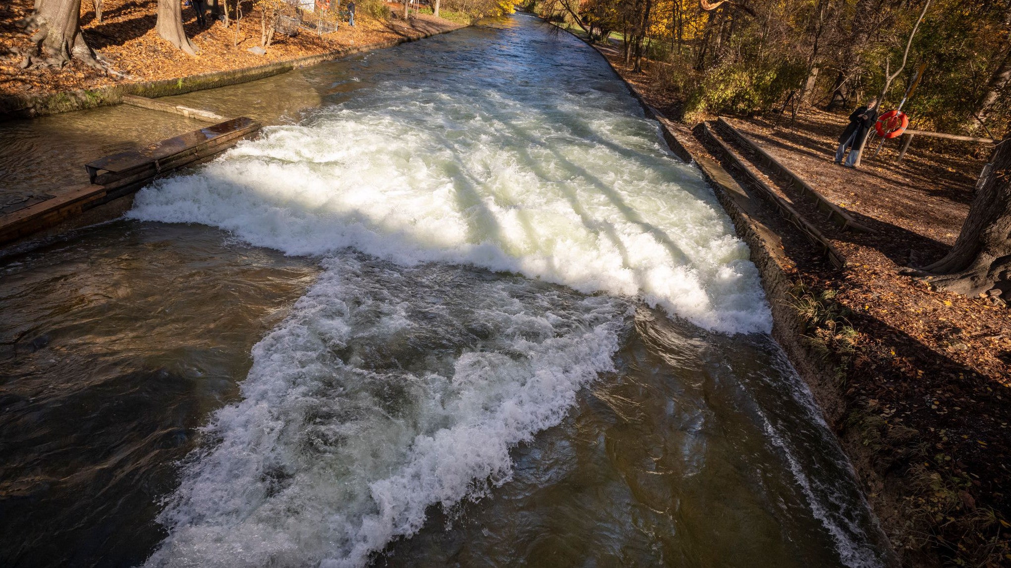 05.11.2025, Bayern, M&uuml;nchen: Passanten stehen am Rande des Eisbachs im Englischen Garten, an der Stelle, an der sich sich normalerweise die Eisbachwelle bildet und die aktuell f&uuml;r den Freizeitsport nicht geeignet ist. Die Eisbachwelle in M&uuml;nchen soll repariert werden. (Peter Kneffel/dpa)