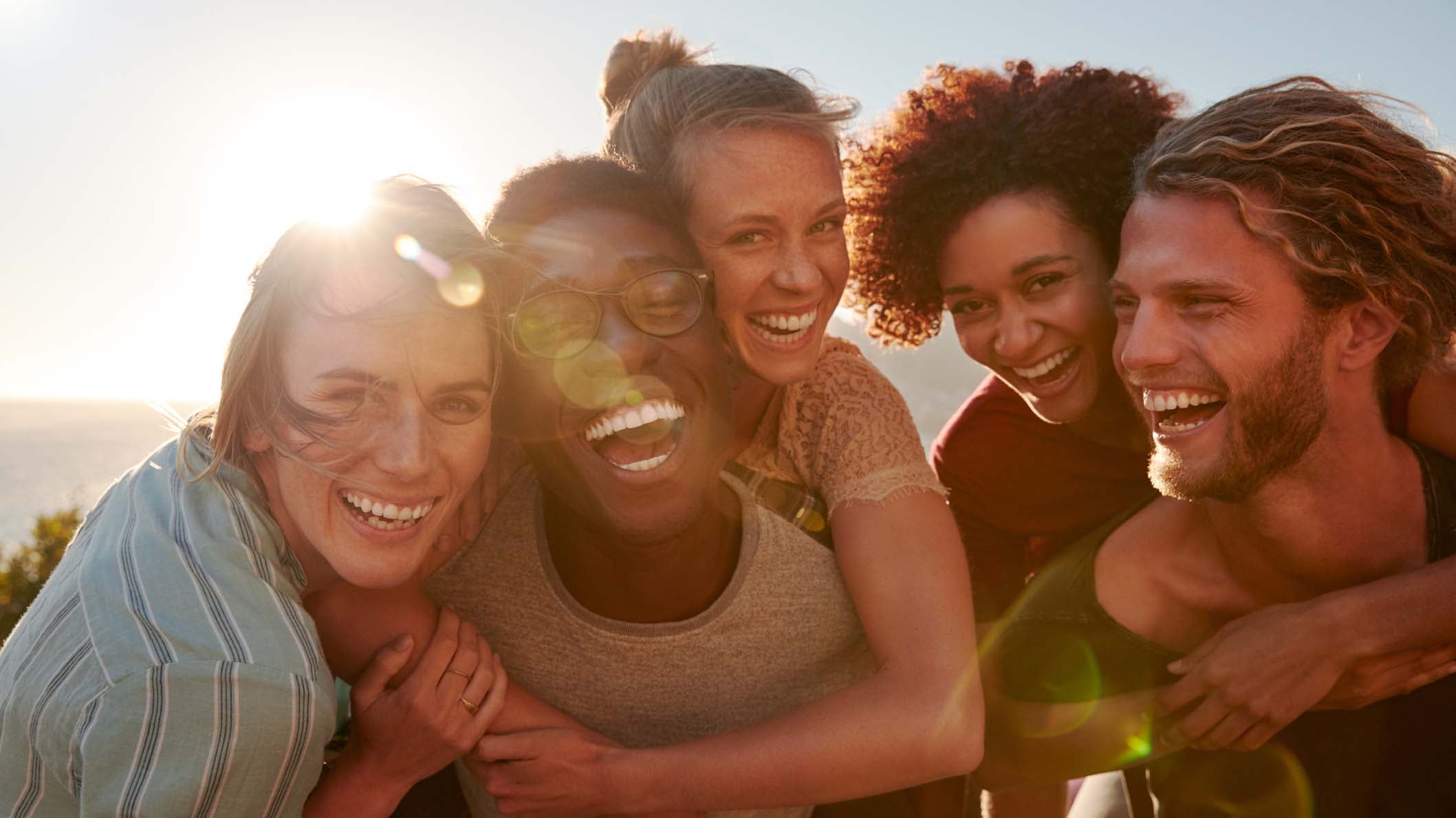 Five millennial friends on a road trip have fun piggybacking at the roadside, front view, lens flare