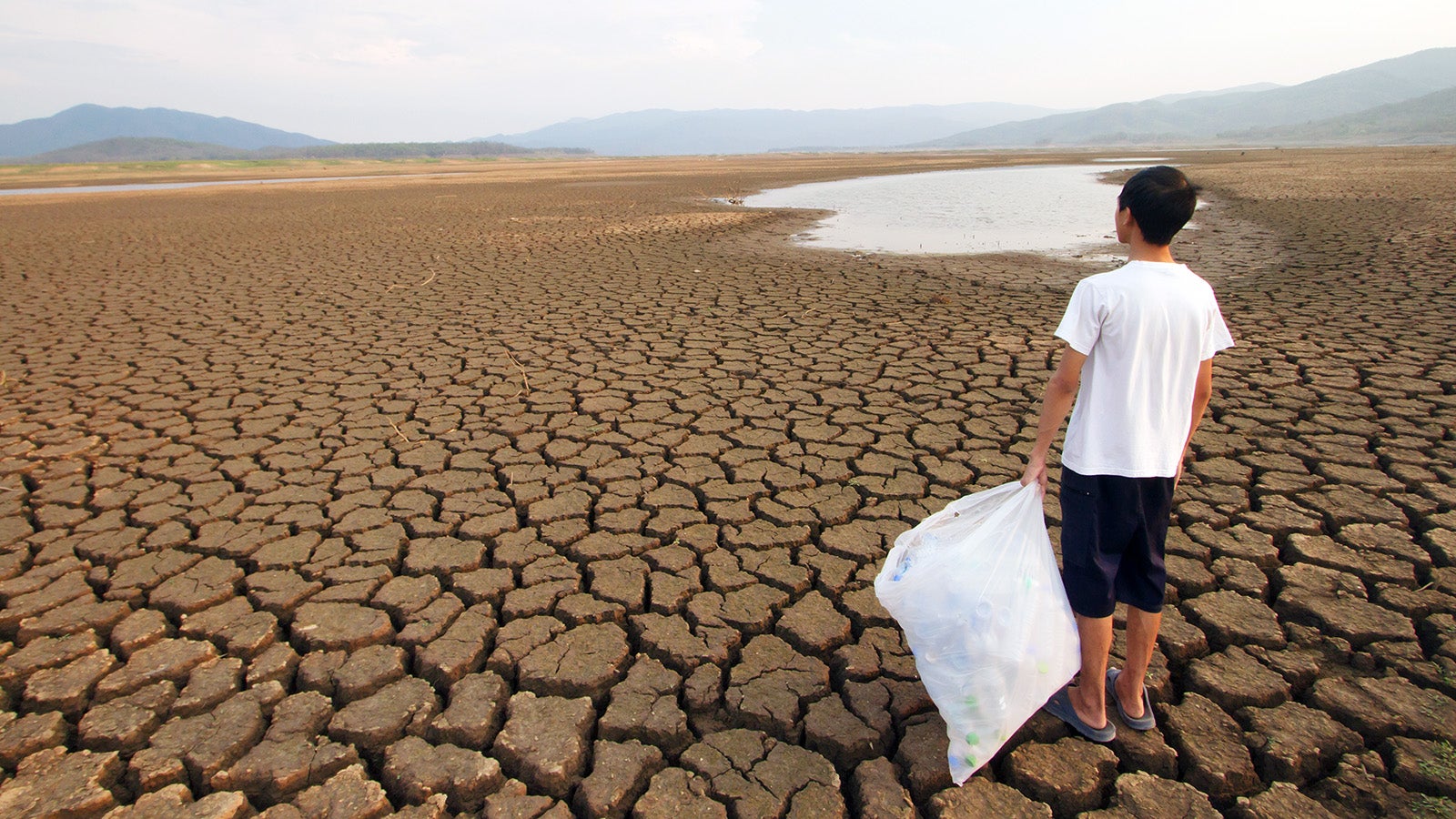Young man holding full plactic bags while looking to the drying lake metaphor Drought and Plastic pollution in the water