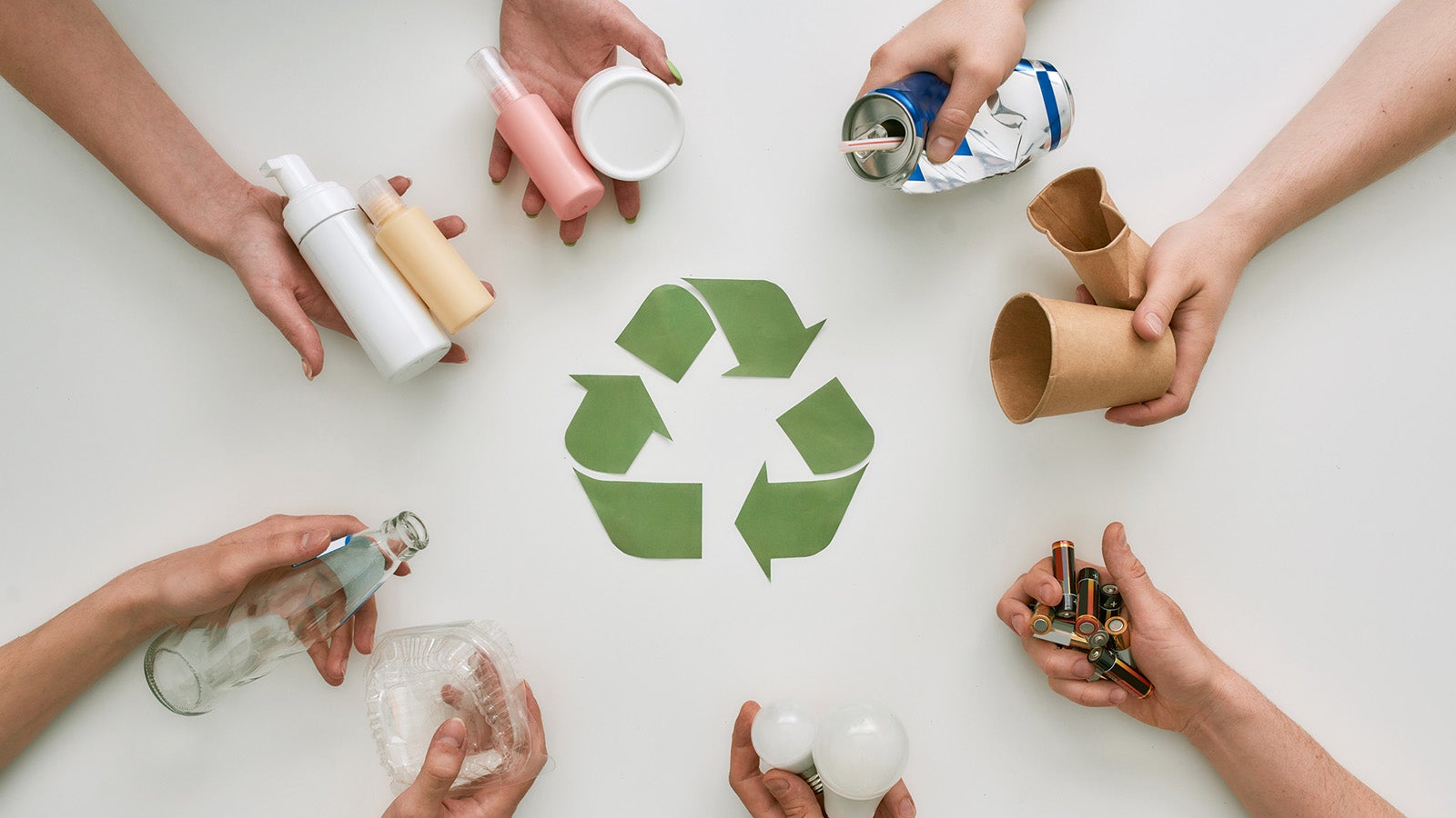 Top view of many hands holding different waste, garbage types with recycling sign made of paper in the center over white background. Sorting, recycling waste concept. Horizontal shot. Top view