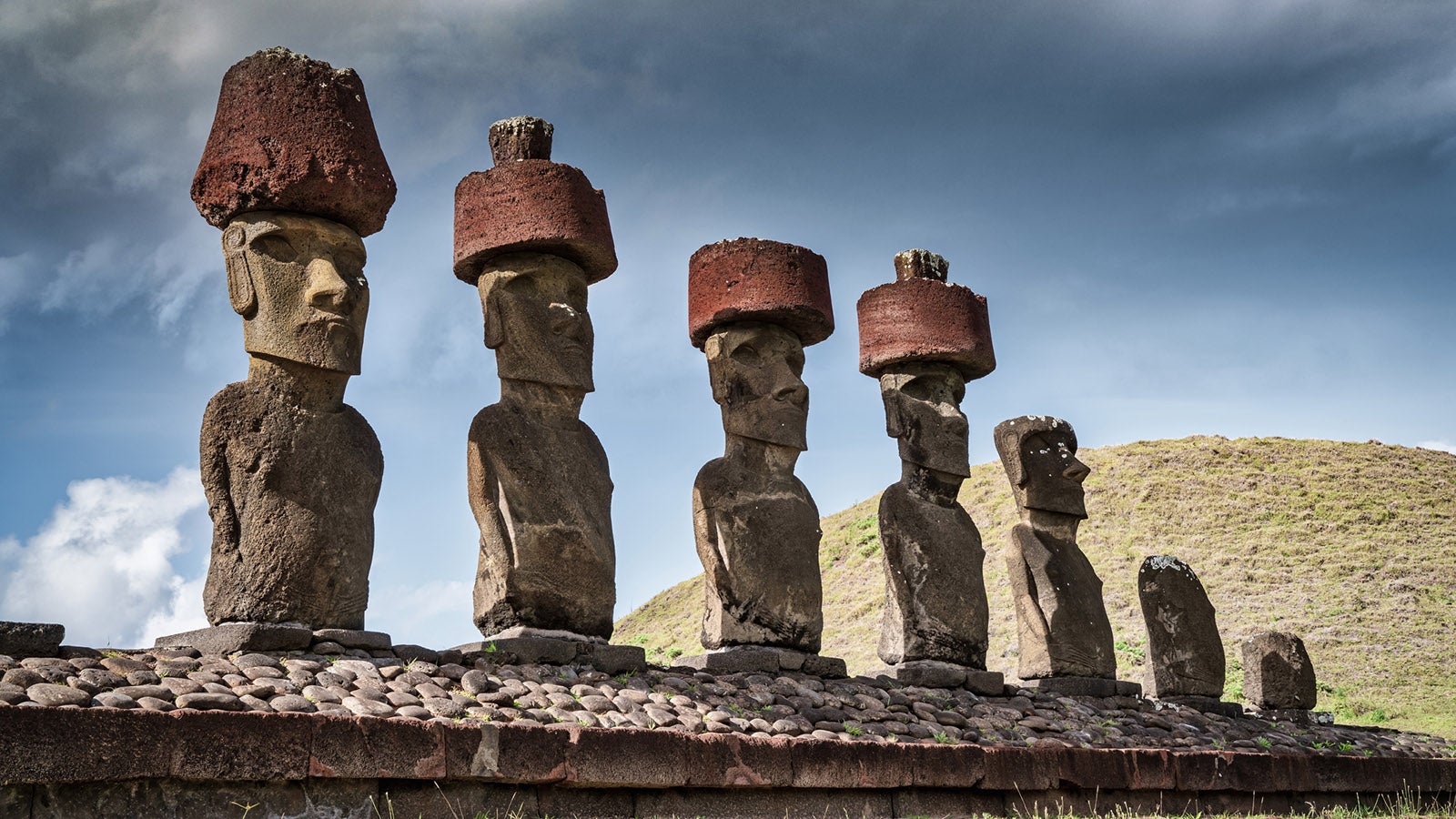 Ahu Nau Nau Ancient Moai Statues, Seven Moais side by side in a row with ancient stone headgear at Anakena Beach under moody summer sky looking towards the beach and pacific ocean. Ahu Nau Nau - Anakena - is also known as Haŋa rau o te 'ariki - The Bay of the King Beach. Anakena Beach, Rapa Nui National Park, Hanga Roa, Easter Island, Isla de Pascua, Polynesia, Oceania, Chile