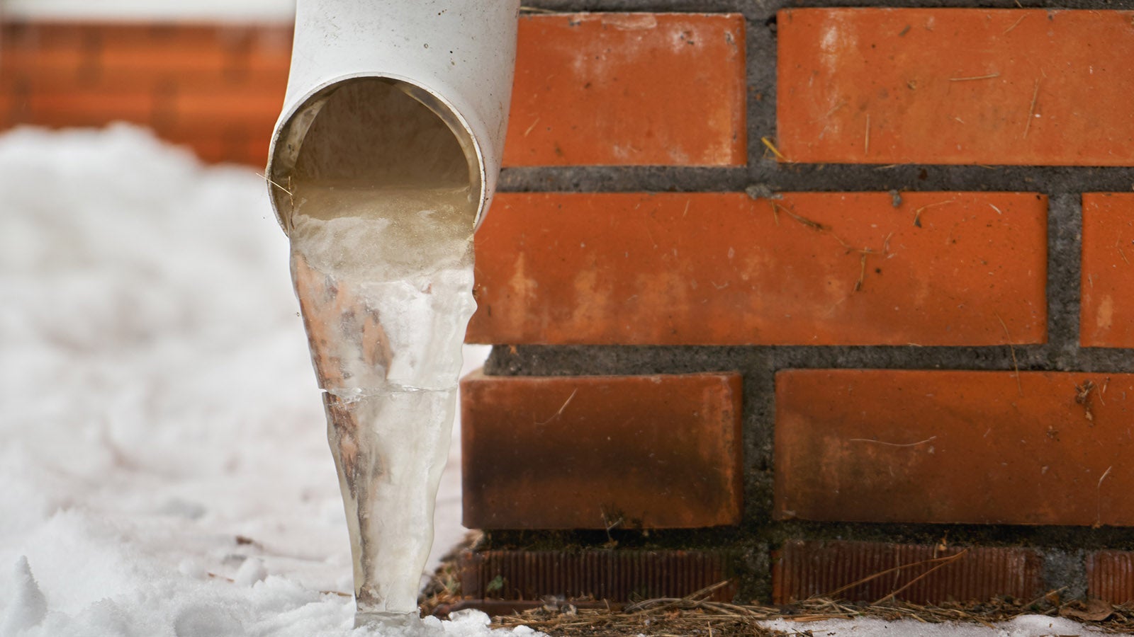 Drain pipe with frozen stream of water near brick wall of a cottage outdoors in winter