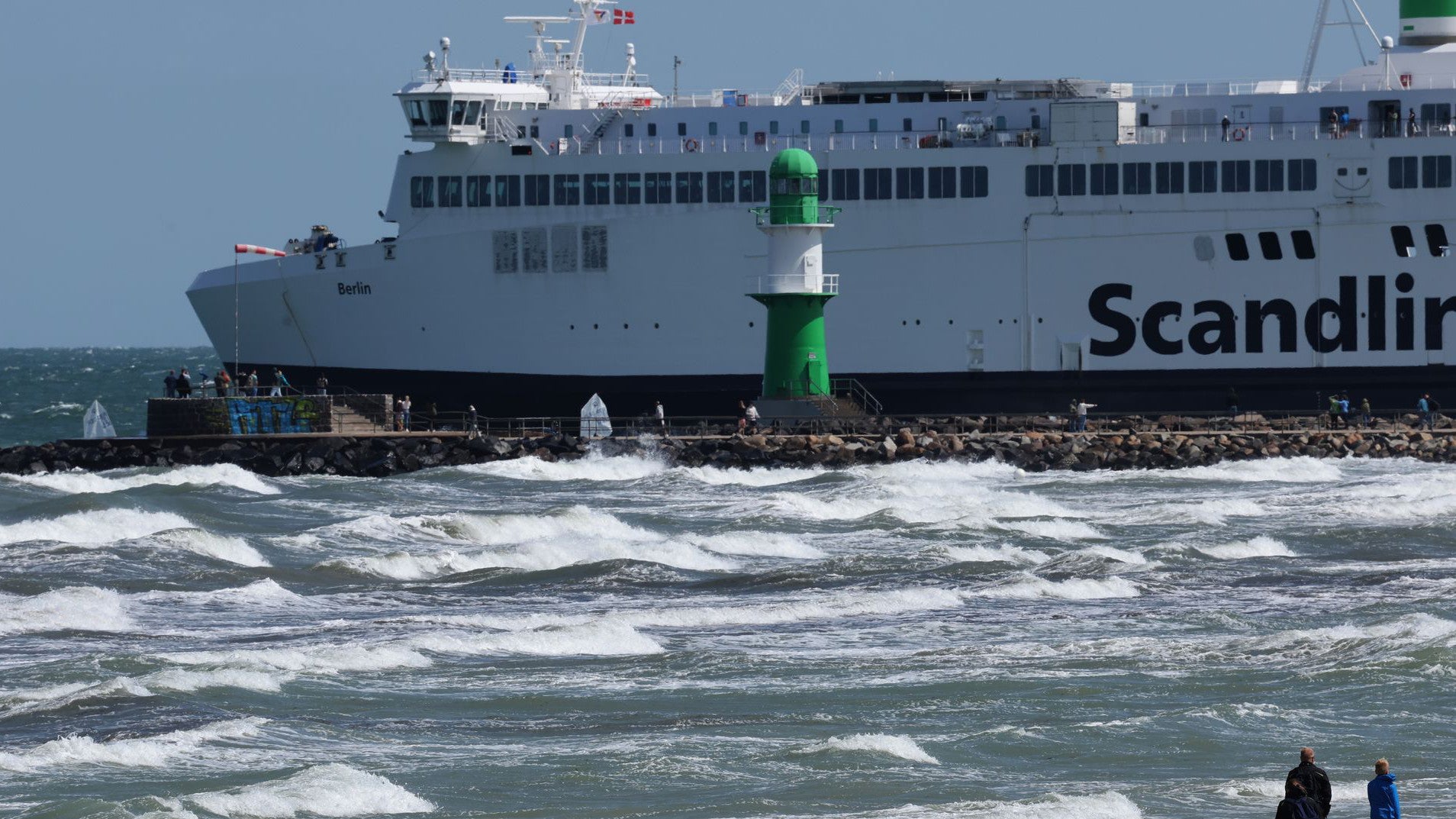 19.06.2025, Mecklenburg-Vorpommern, Warnem&uuml;nde: Spazierg&auml;nger sind an der aufgew&uuml;hlten Ostsee unterwegs, eine Scandlines-F&auml;hre macht sich auf den Weg Richtung D&auml;nemark. Starker Wind hat zu einem Badeverbot gef&uuml;hrt, die Wassersportler freuen sich &uuml;ber das Wetter. (Bernd W&uuml;stneck/dpa)