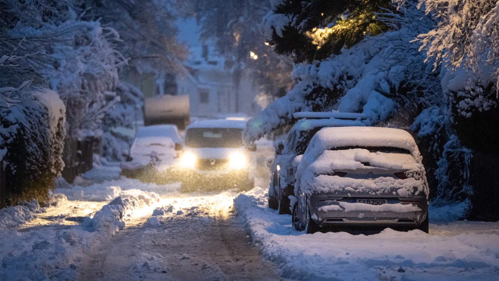 20.02.2026, Bayern, M&uuml;nchen: Ein Transporter f&auml;hrt mit eingeschaltetem Licht durch eine schneebedeckte Wohnstra&szlig;e, w&auml;hrend parkende Fahrzeuge unter einer dichten Schneeschicht stehen. Anhaltende Schneef&auml;lle sorgen f&uuml;r winterliche Stra&szlig;enverh&auml;ltnisse und erschweren den Verkehr. Foto: Lukas Barth-Tuttas/dpa +++ dpa-Bildfunk +++