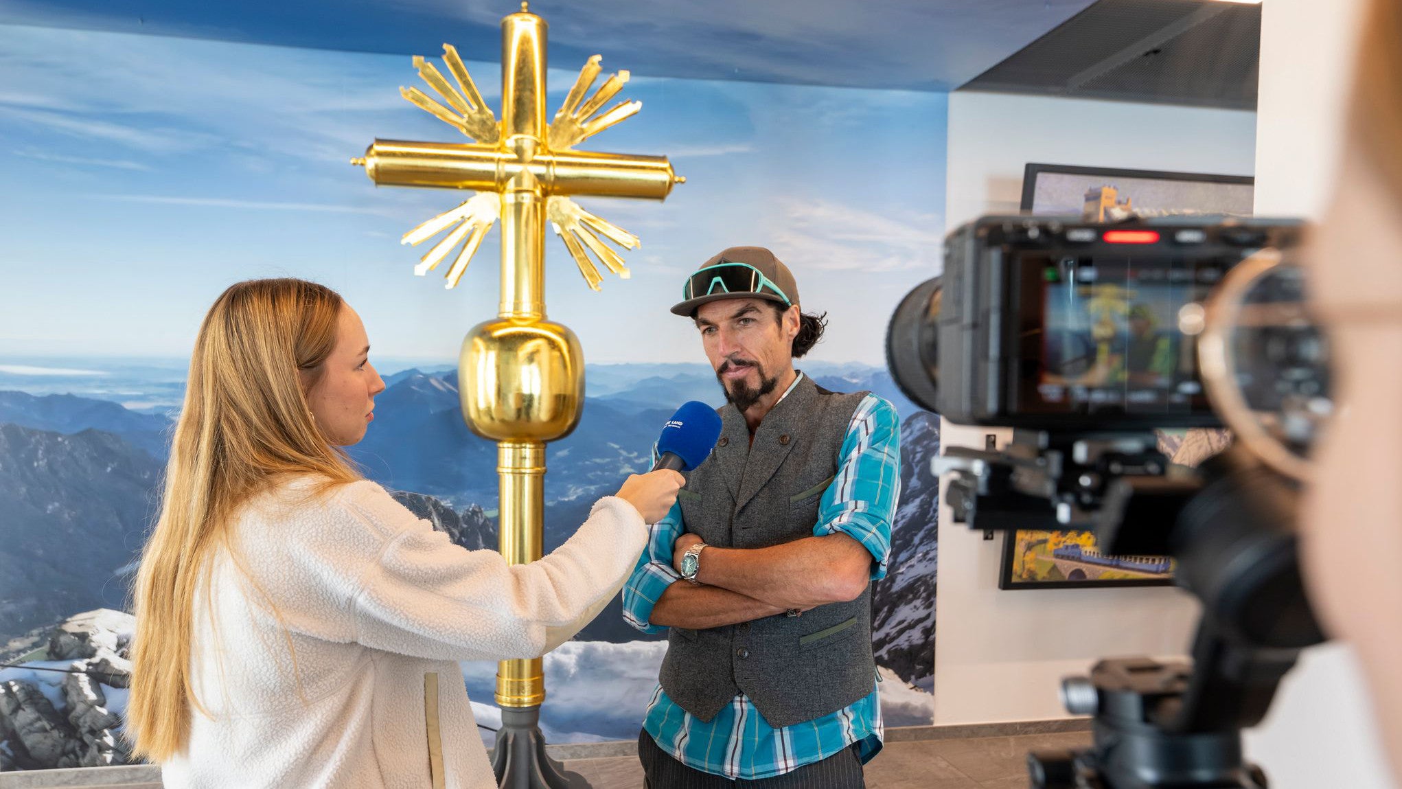 03.07.2025, Bayern, Garmisch-Partenkirchen: Der K&uuml;nstler Bernhard Rieger wird vor dem zweiten Gipfelkreuz auf der Zugspitze interviewt. Das goldene Gipfelkreuz an der Zugspitze ist mit Hunderten Stickern beklebt, mit denen sich Besucher auf Deutschlands h&ouml;chstem Berg verewigt haben. Nun muss es restauriert werden. (Peter Kneffel/dpa)
