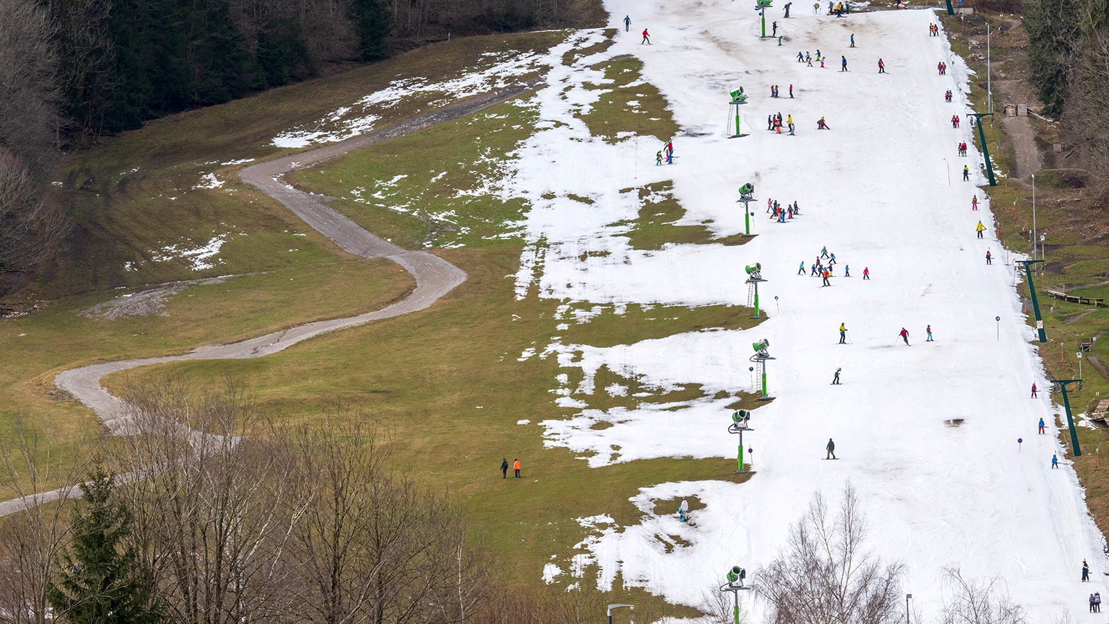 ARCHIV - 04.01.2024, Bayern, Brauneck: Skifahrer nutzen am Vormittag die beschneite Fl&auml;che im Talbereich f&uuml;r ihr k&uuml;hles Wintervergn&uuml;gen. Links zieht sich ein Feldweg durch eine gr&uuml;ne Wiese. (zu dpa: &laquo;Bayerns Skigebiete r&uuml;sten sich f&uuml;r die Saison&raquo;) Foto: Peter Kneffel/dpa +++ dpa-Bildfunk +++