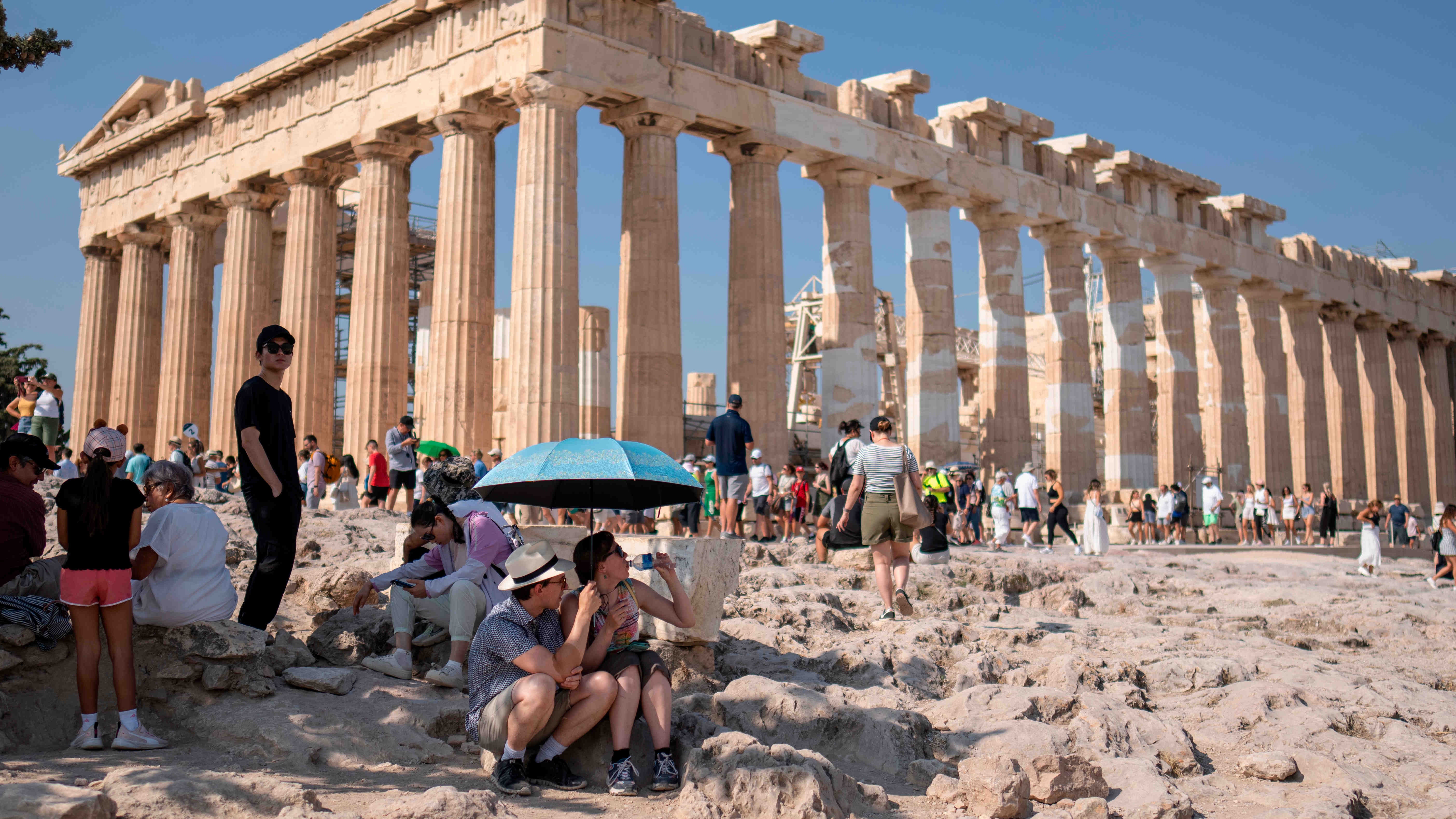 dpatopbilder - 13.07.2023, Griechenland, Athen: Eine Touristin trinkt Wasser, w&auml;hrend sie und ein Mann unter einem Regenschirm vor dem Parthenon-Tempel auf dem Akropolis-H&uuml;gel Schutz vor der Sonne suchen. Am Wochenende werden in Griechenland und auf Zypern, aber auch im Westen der T&uuml;rkei H&ouml;chsttemperaturen von &uuml;ber 40 Grad erwartet. Foto: Petros Giannakouris/AP/dpa +++ dpa-Bildfunk +++