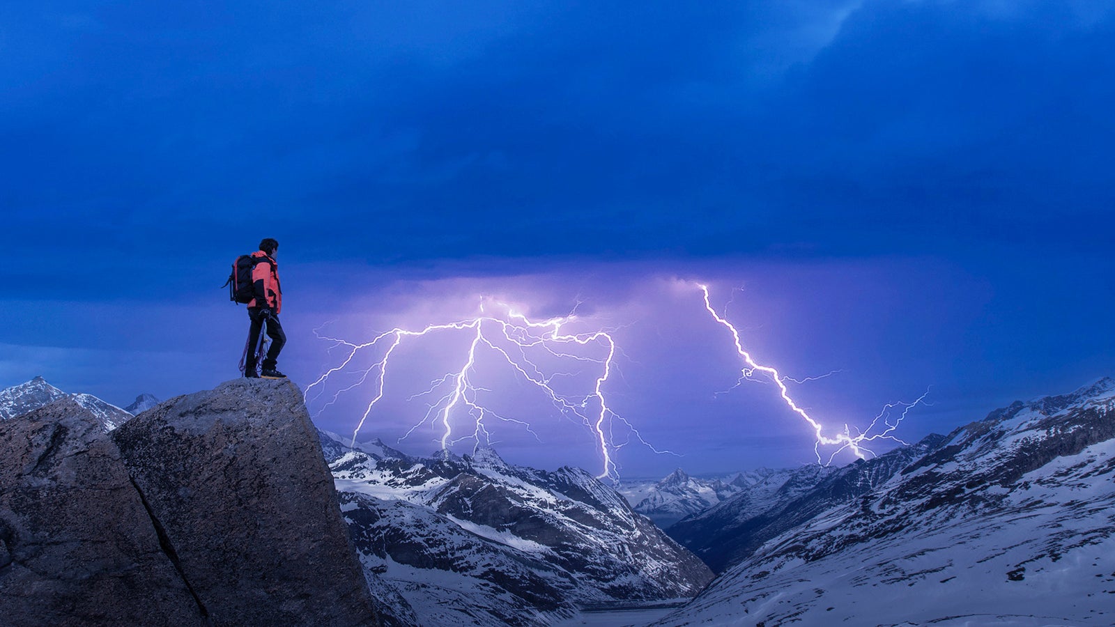 Side view of a caucasian climber standing on a rock at night watching a a thunderstorm with lightning over a mountain range.