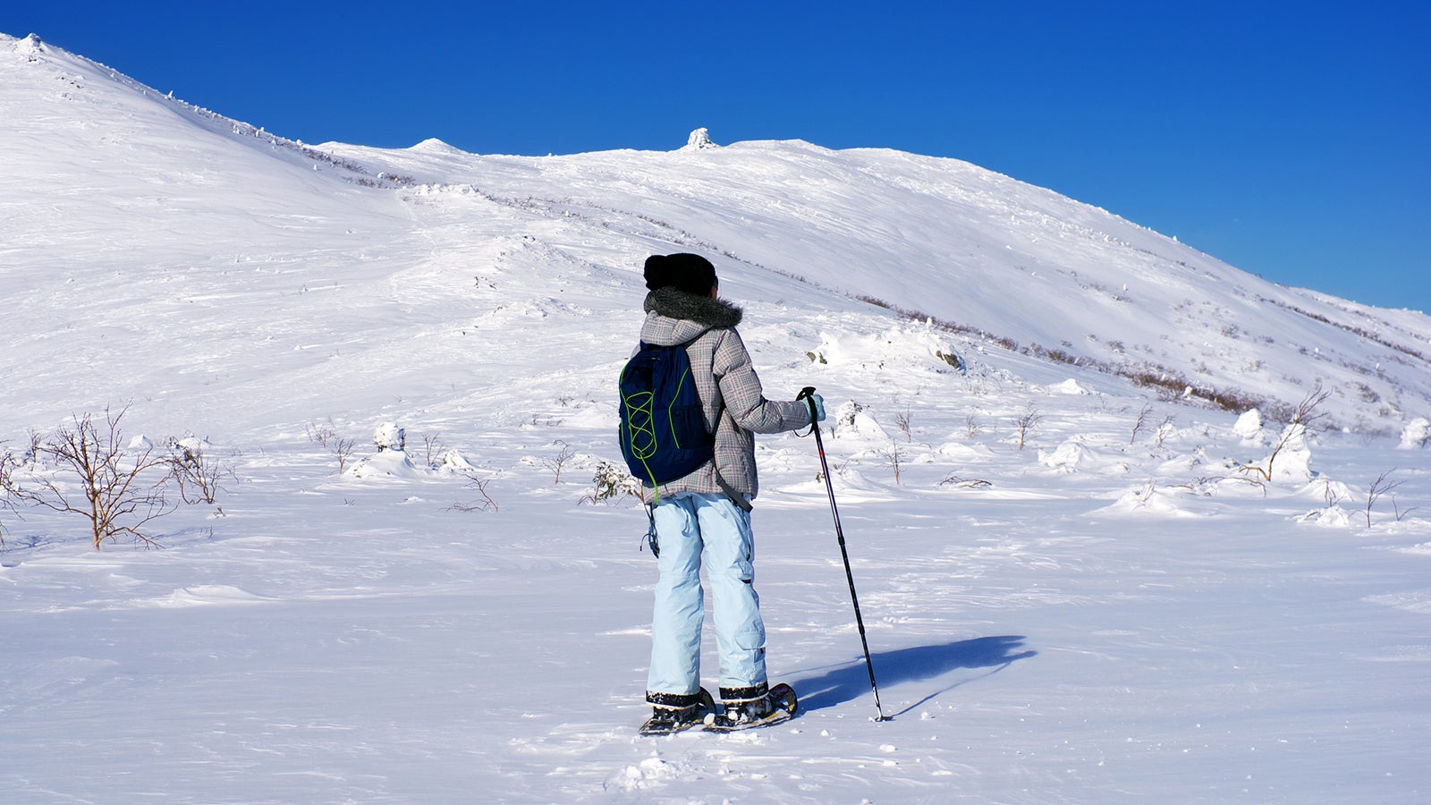 Young woman backpacker snowshoeing winter mountain, ski poles. Blue sky, white snow peak.