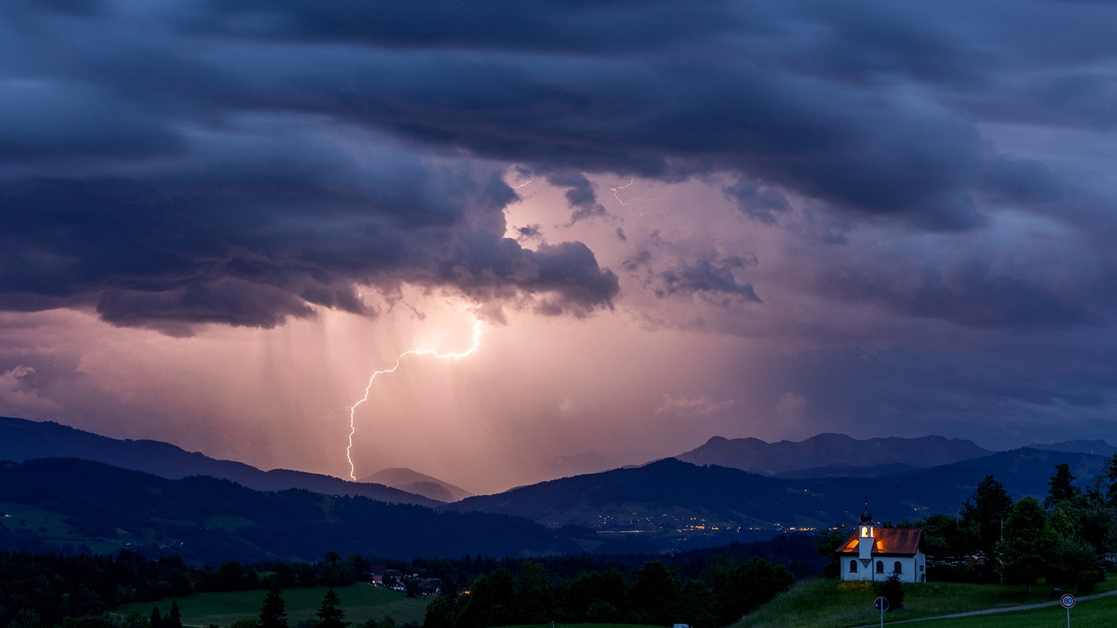 Blitze entladen sich &uuml;ber den Alpen w&auml;hrend eines aufziehenden Gewitters am Abend hinter der St. Hubertus Kapelle in Scheidegg. Foto: Jan Eifert/dpa