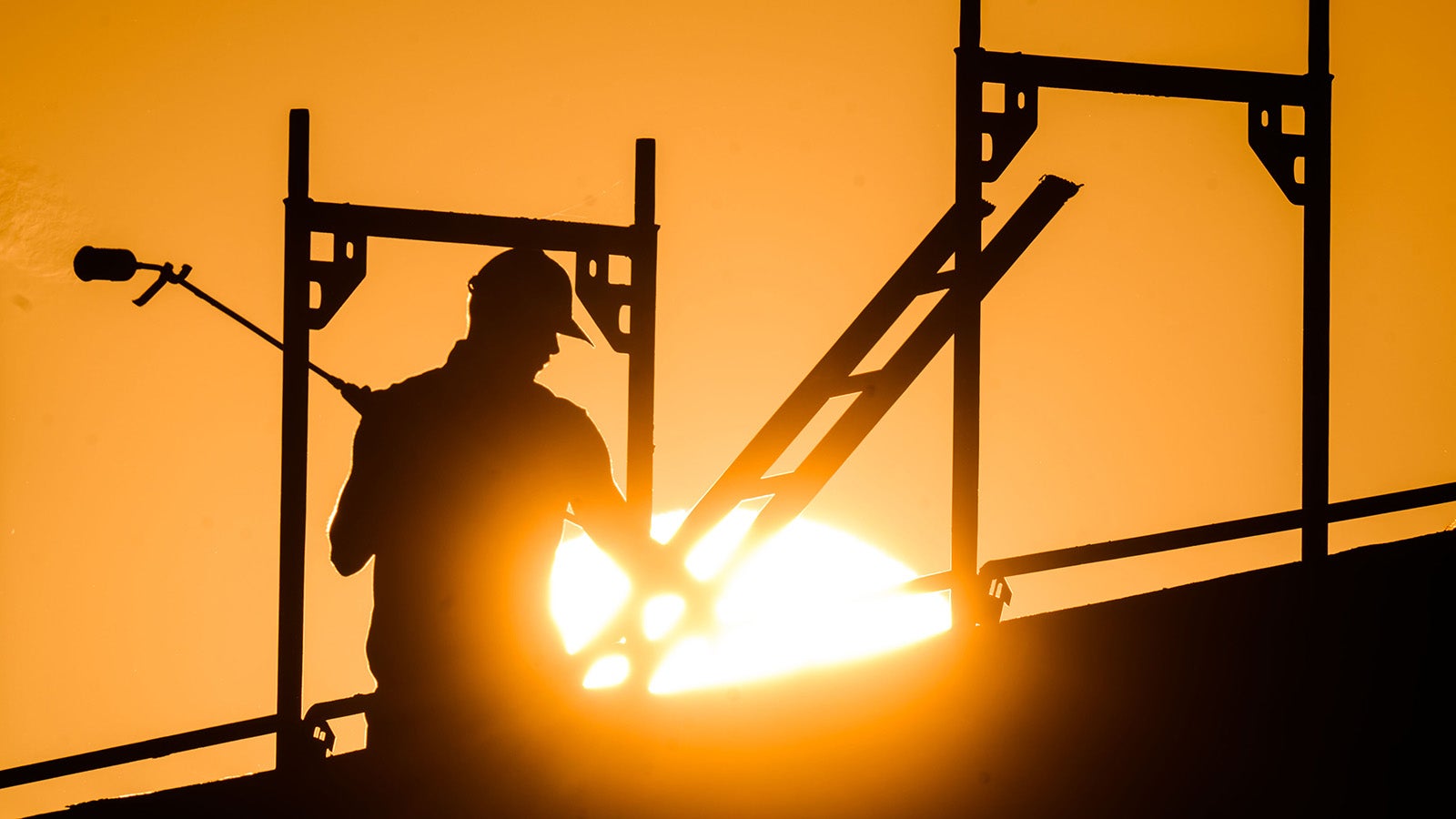 Silhouette einer Person mit Bauhelm und Werkzeug auf einem Ger&uuml;st vor tiefstehender Sonne. dpa