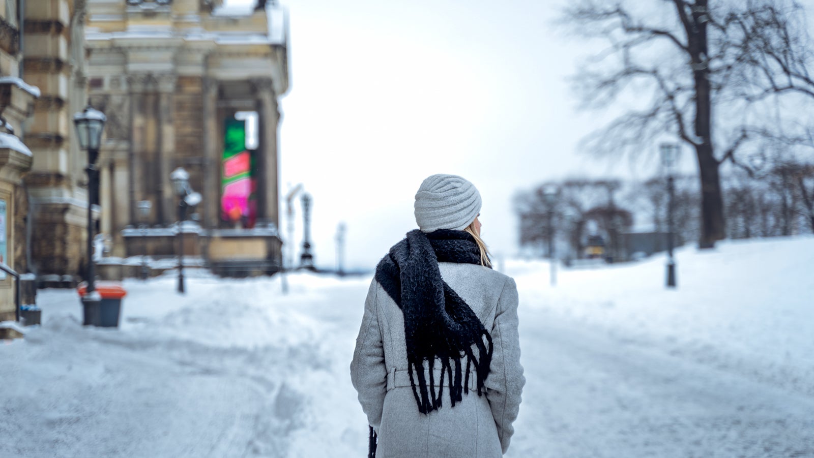 Back view of anonymous female in coat with scarf and hat holding hands in pockets and looking away while strolling on snowy road on winter day in Dresden, Germany