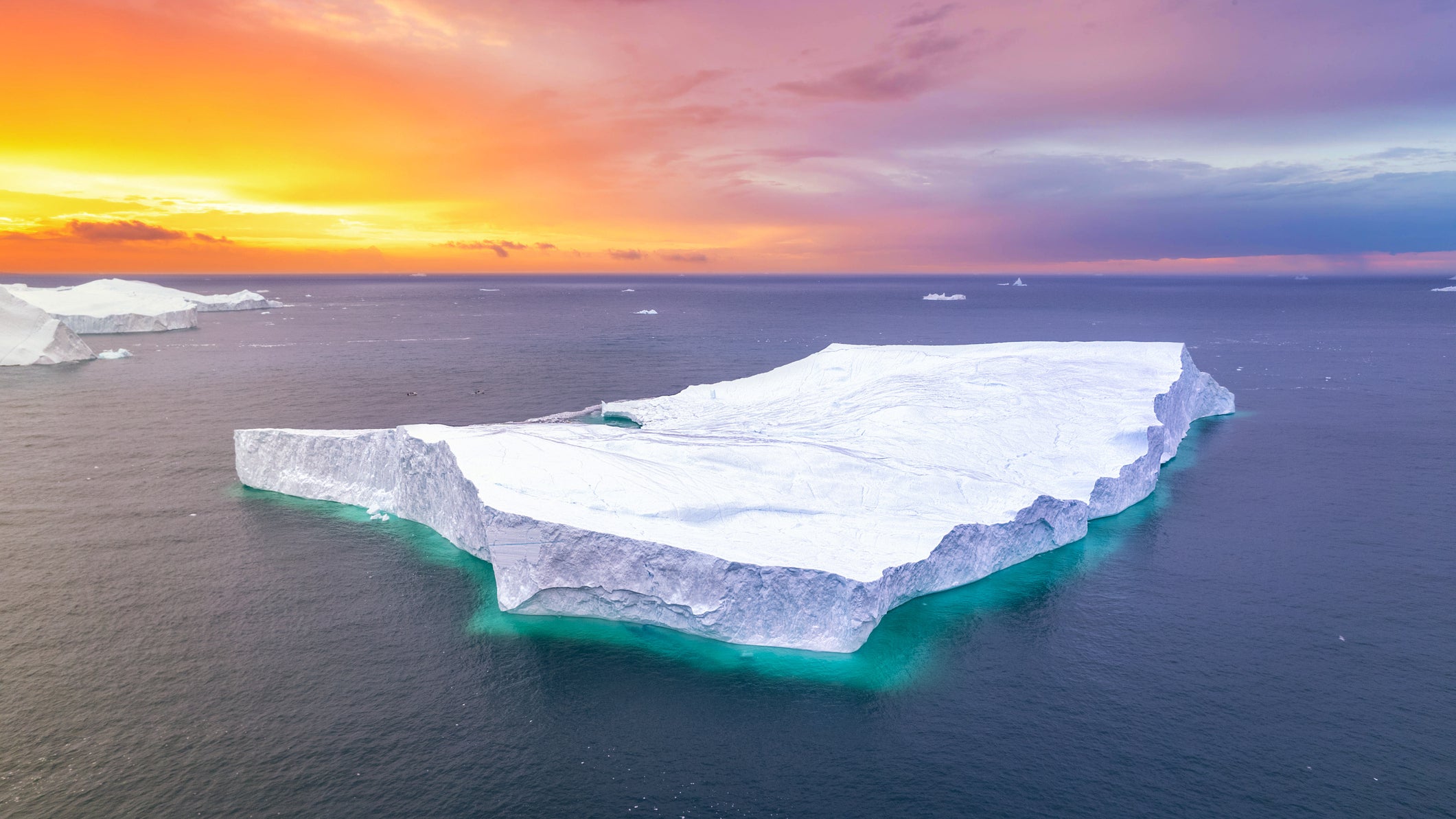 Majestic aerial view of a large tabular iceberg drifting in the dark Arctic waters near Ilulissat during the vibrant midnight sun, concept of global warming and climate change. The glowing sky in shades of orange, pink, and purple contrasts beautifully with the icy blue tones of the floating glacier. This striking image captures the stark beauty of the polar environment and highlights the impact of climate change on the fragile Arctic ecosystem. It powerfully represents the grandeur and vulnerability of nature in a warming world. Ilulissat, Greenland