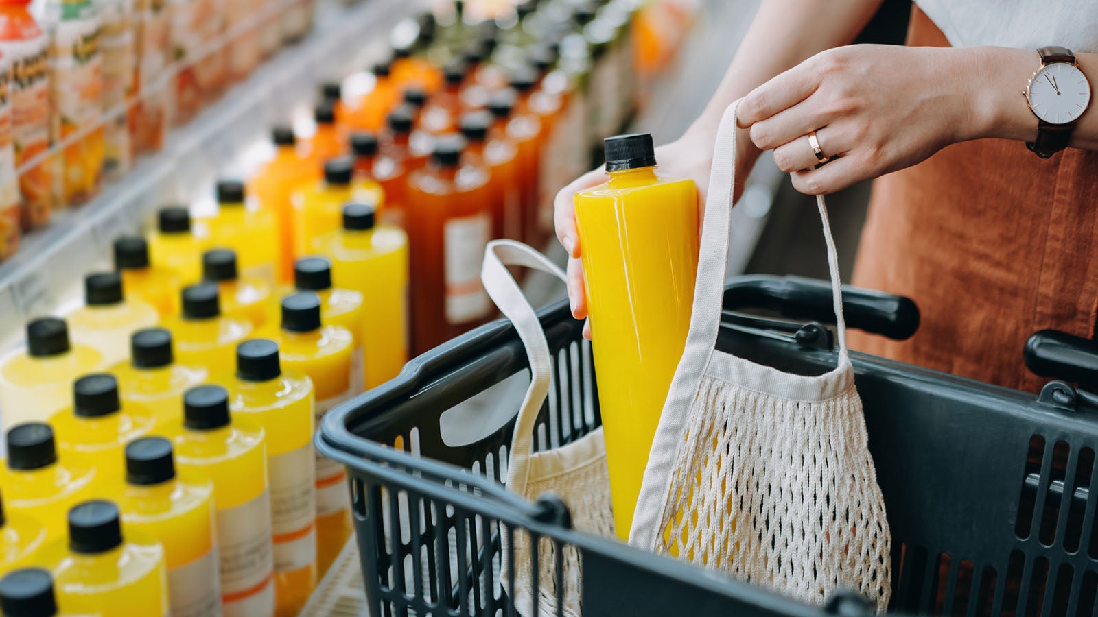 Cropped shot of young Asian woman shopping for fresh fruit juice from refrigerated shelves in supermarket and putting a bottle of fresh squeezed orange juice into cotton mesh eco bag in a shopping cart. Zero waste concept